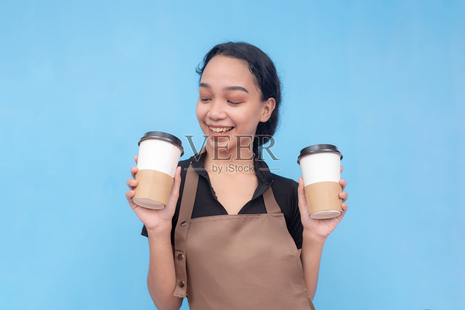一位年轻友好的亚洲女咖啡师拿着两杯咖啡，感到非常幸福。穿着棕色围裙和黑色衬衫，背景是浅蓝色照片摄影图片