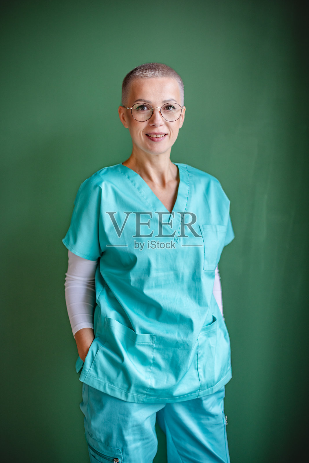 身穿医疗工作服的女性医护人员，双手插在口袋里，面带微笑地站在医疗机构前的绿色背景前照片摄影图片