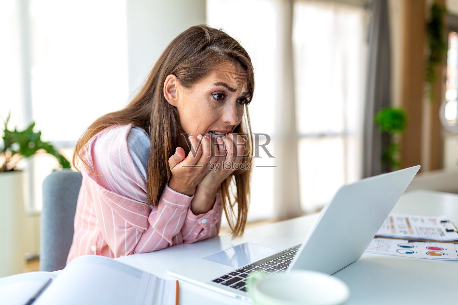 焦虑和工作压力，年轻女子咬指甲，有心理健康问题的人，医疗保健和医学概念。一位迷人的女子坐在办公桌前，看着笔记本电脑，表情惊恐。照片摄影图片