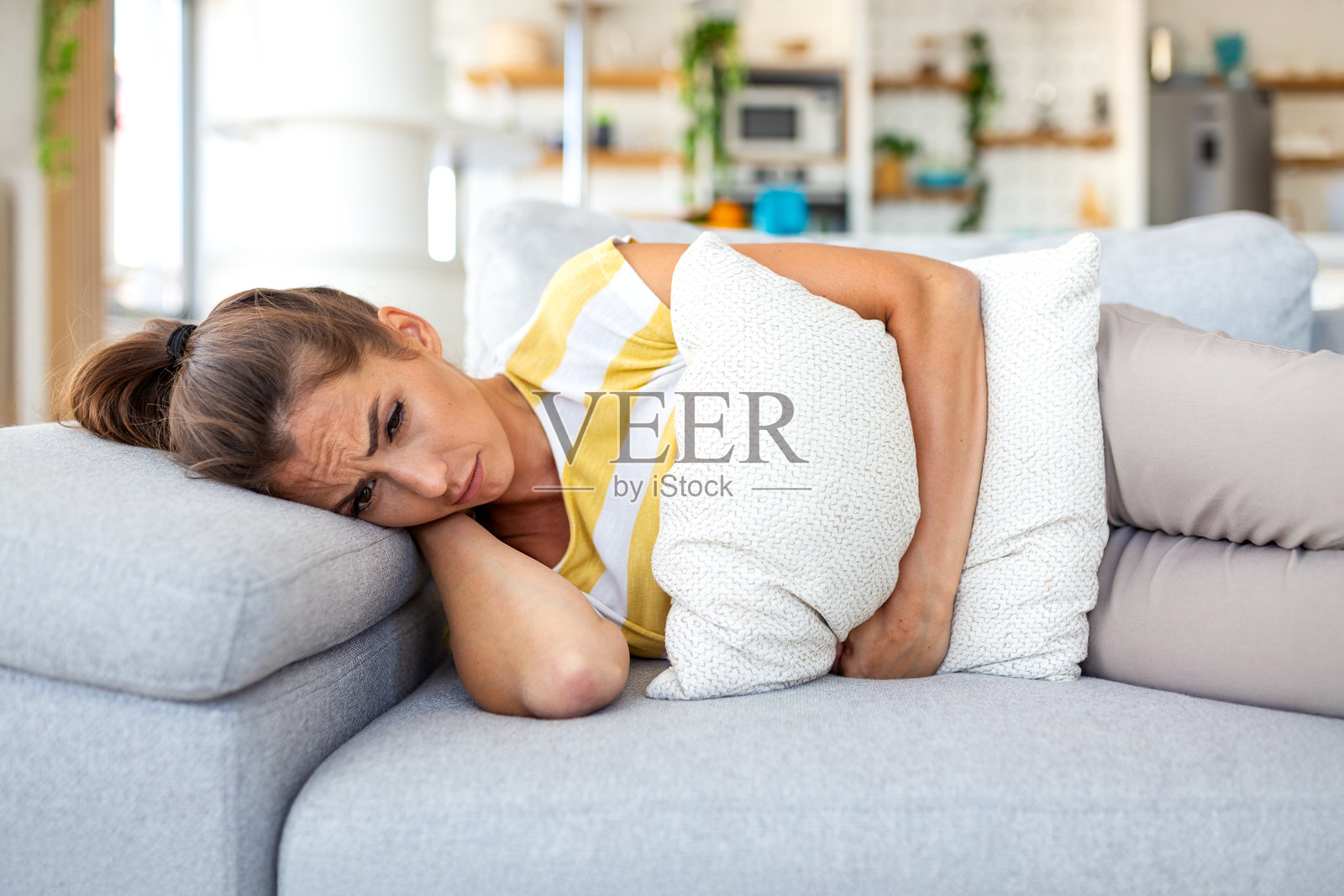 悲伤的女性躺在沙发上，腹痛难忍，抱着枕头。经期痉挛、腹部疼痛和孤独感概念。年轻女性在家中遭受经期不适的痛苦。医疗保健与医药。照片摄影图片