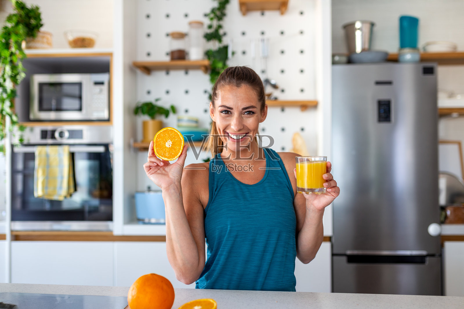 快乐运动的女性在厨房里拿着橙子和一杯新鲜果汁。健康生活方式、营养和养生理念。锻炼后，美丽的女孩在家中带着维生素饮料微笑着。照片摄影图片