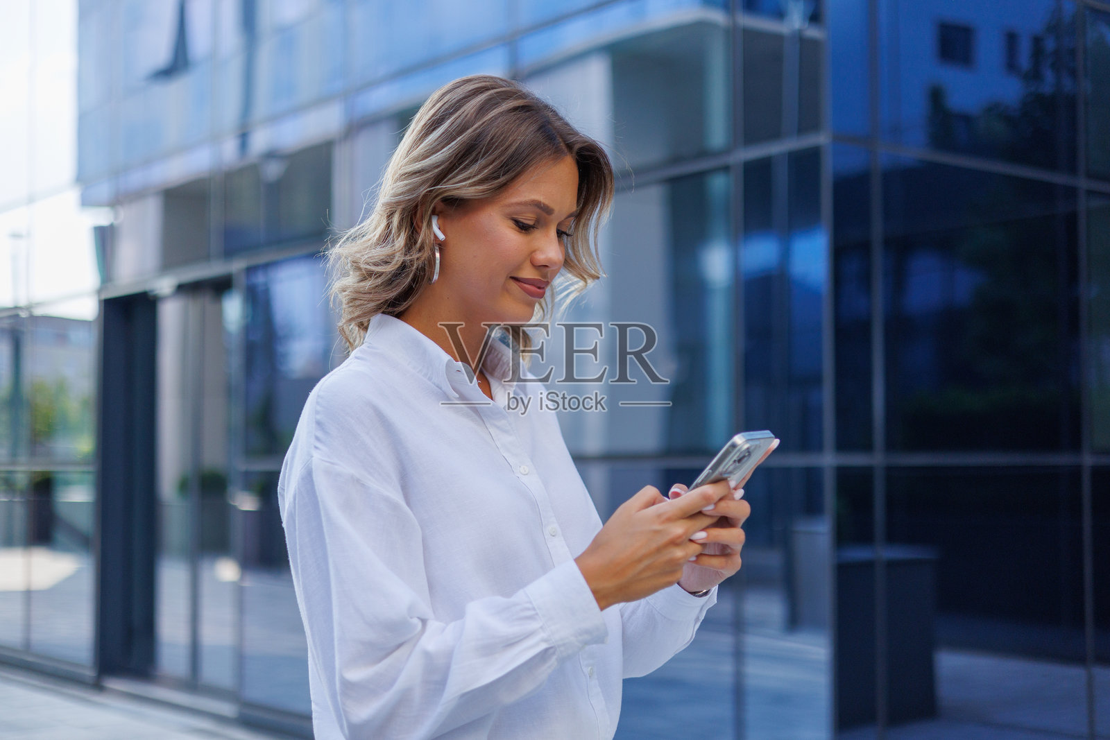 年轻的女商人使用智能手机和无线耳机，在现代办公楼附近行走照片摄影图片