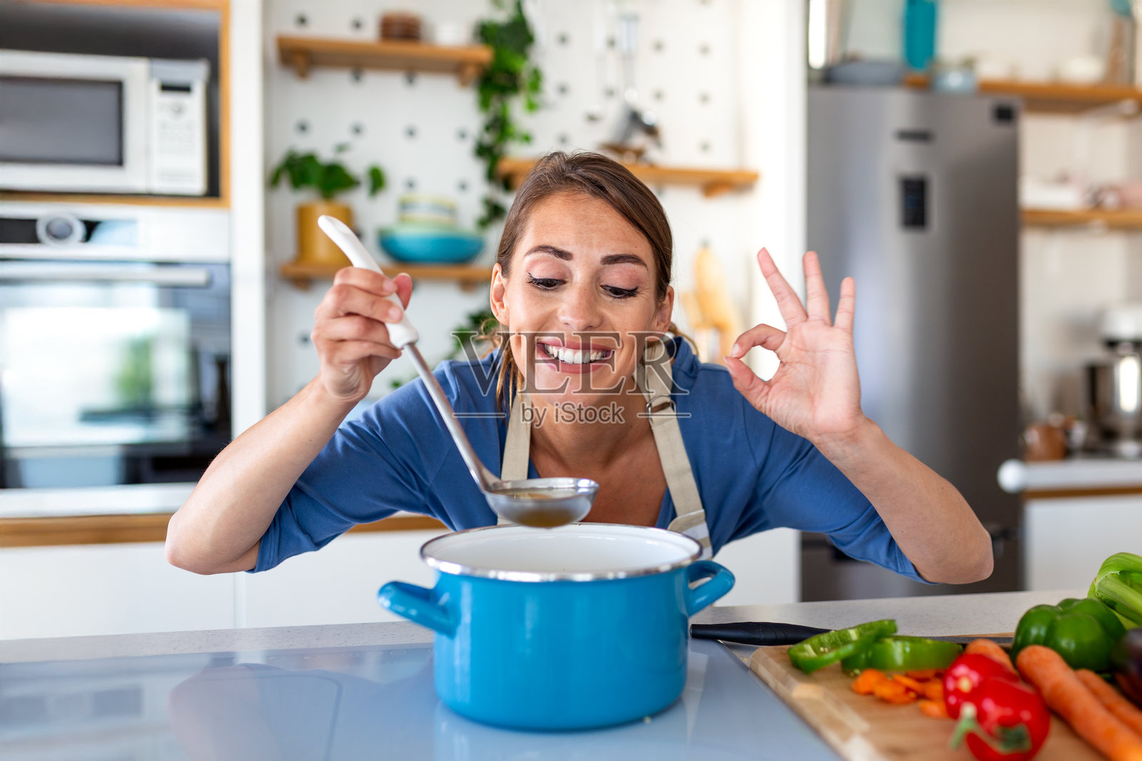 穿着围裙的快乐女子在厨房里，一边用勺子品尝蓝色锅里的汤，一边竖起大拇指。微笑的女厨师正在享用自制的餐点。健康烹饪、居家生活和成功的概念。照片摄影图片