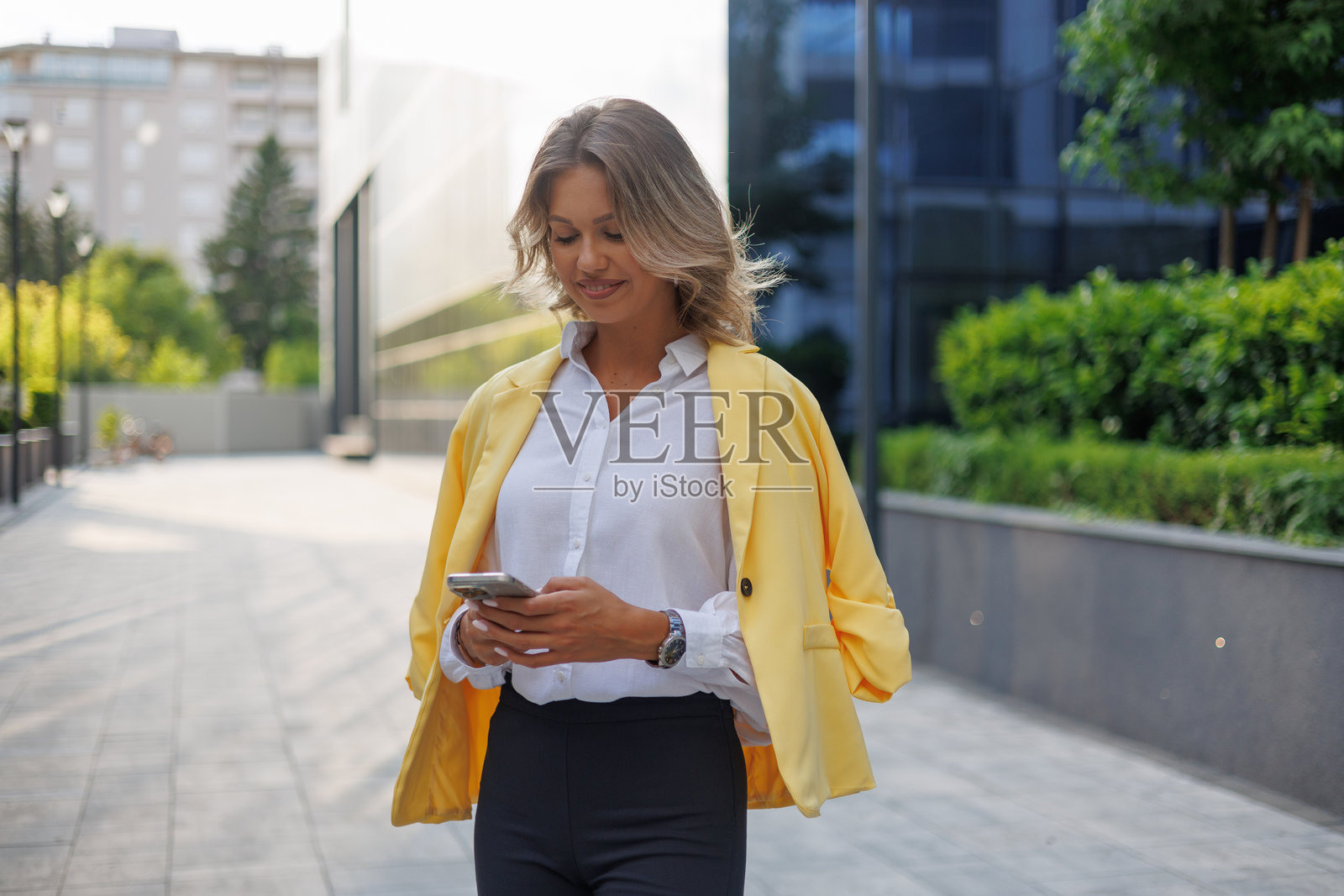 一位年轻女子穿着黄色西装外套，在城市环境中边走边查看智能手机照片摄影图片