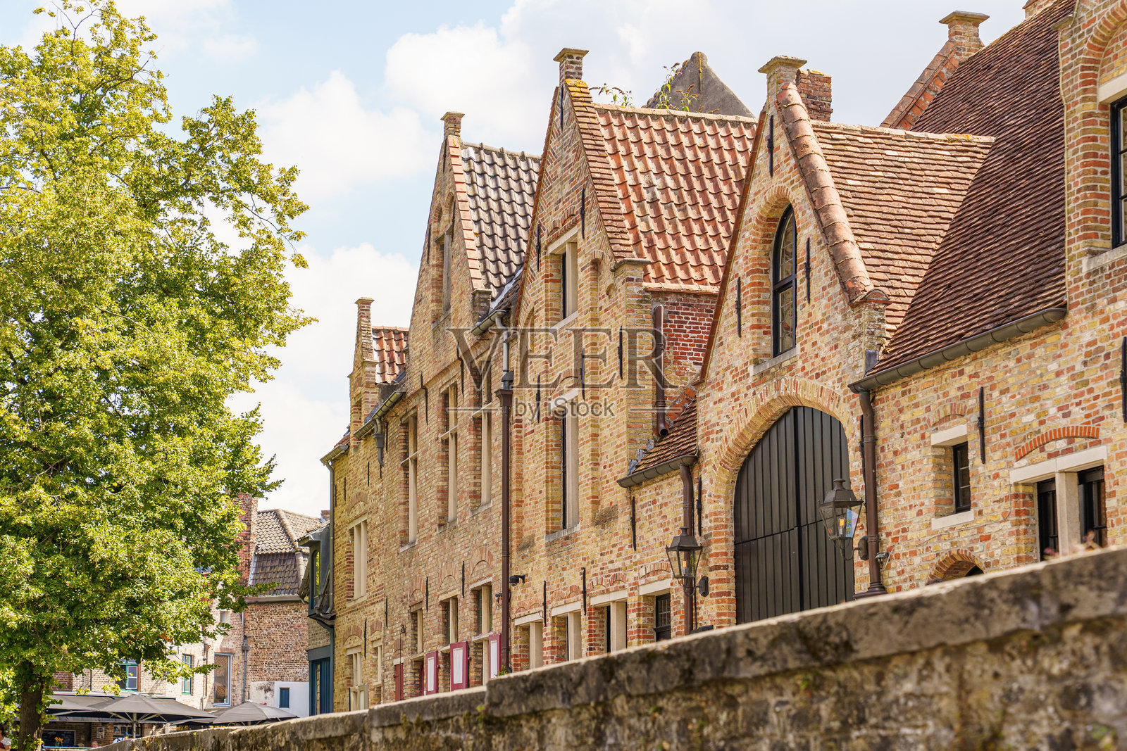 比利时布鲁日河畔的红砖房，有尖尖的屋顶，阳光明媚，画面左右两侧有绿植。照片摄影图片