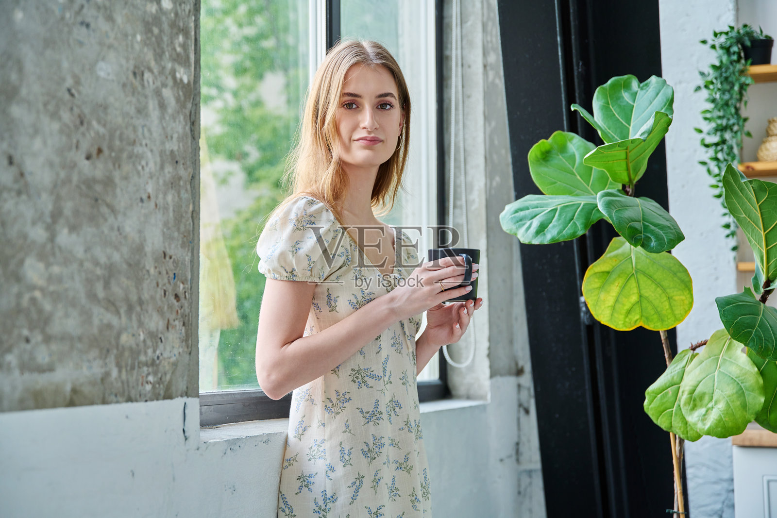 年轻美丽的微笑浪漫女孩，手持杯子看向镜头，站在窗边照片摄影图片