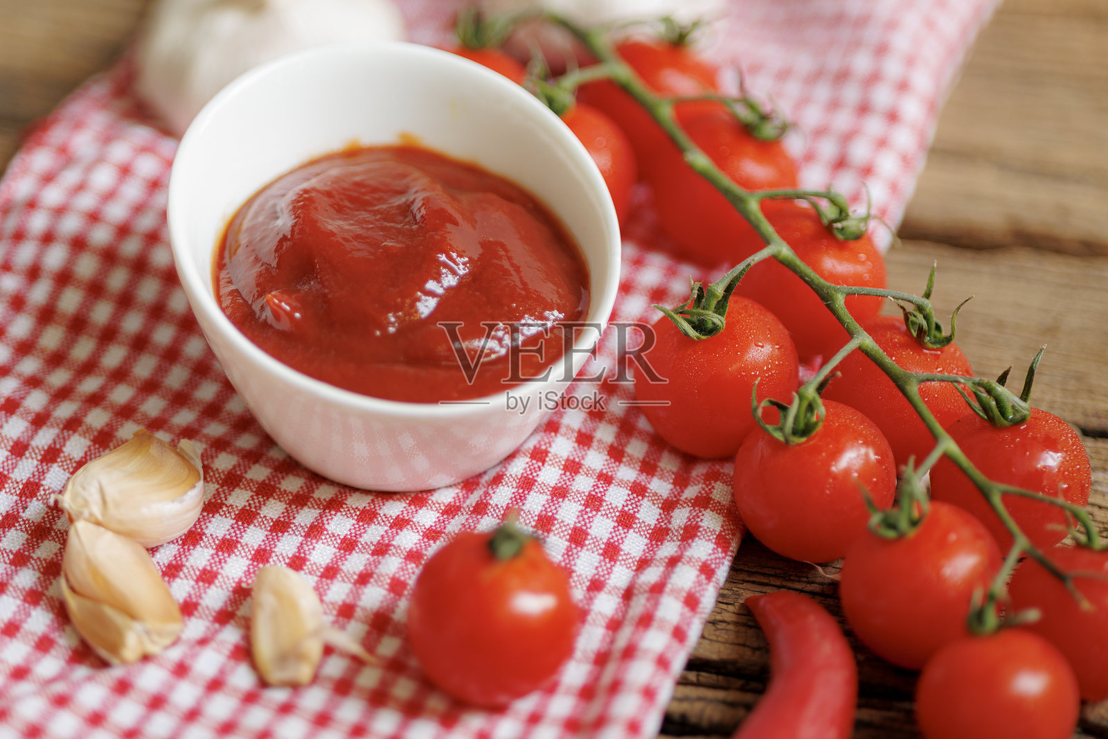 一碗浓郁的番茄酱放在红白格子上，周围散落着新鲜的樱桃番茄、大蒜瓣和辣椒，营造出质朴诱人的场景照片摄影图片