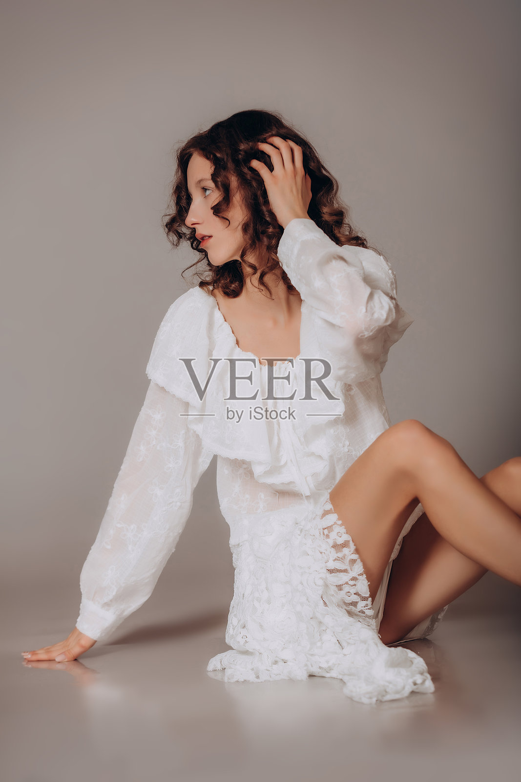 一位年轻美丽的棕发卷发女孩，身穿白色衬衫和蕾丝裙，站在灰色背景前照片摄影图片