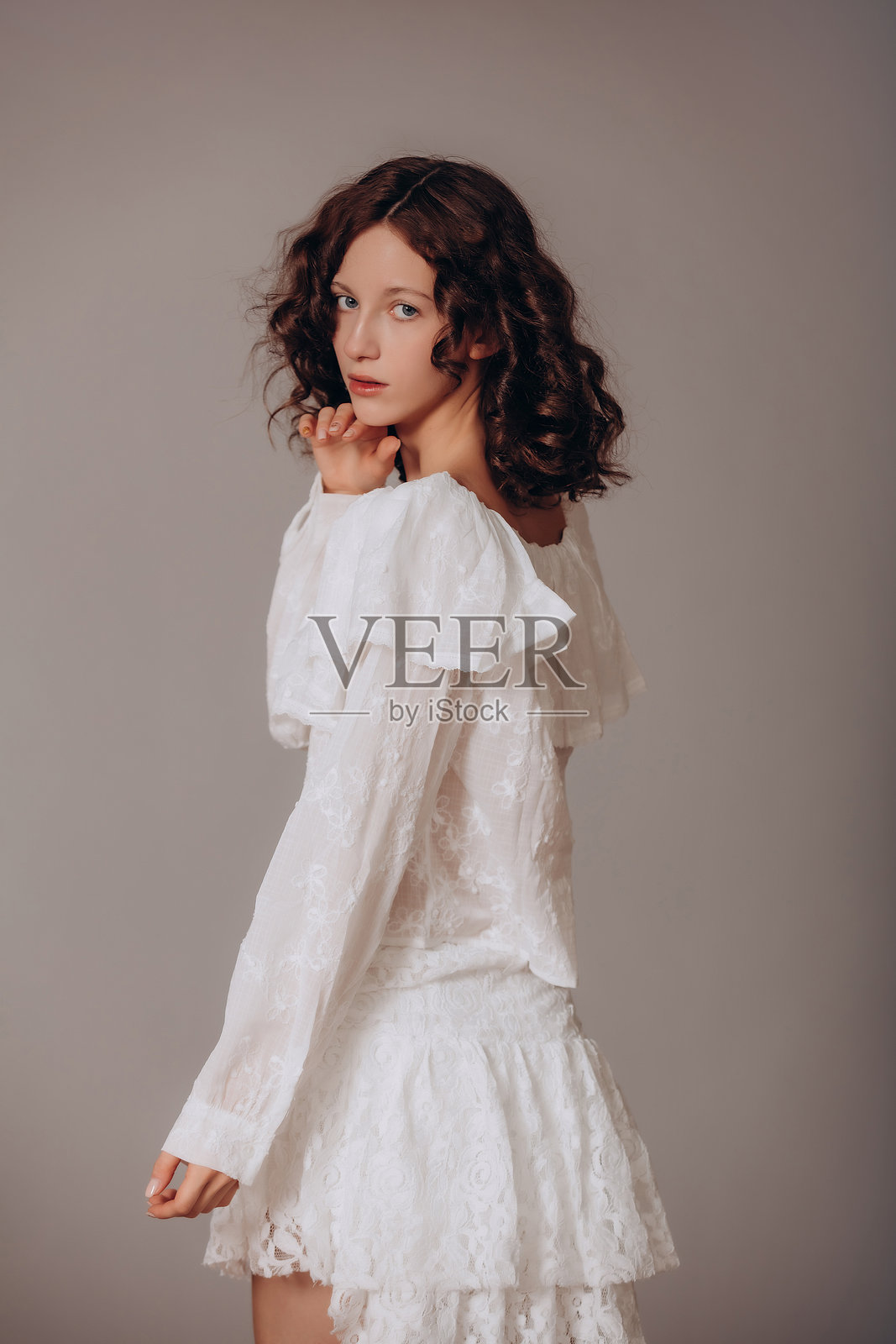 模特的年轻美丽的棕发卷发女孩，身穿白色衬衫和蕾丝裙，站在灰色背景前照片摄影图片