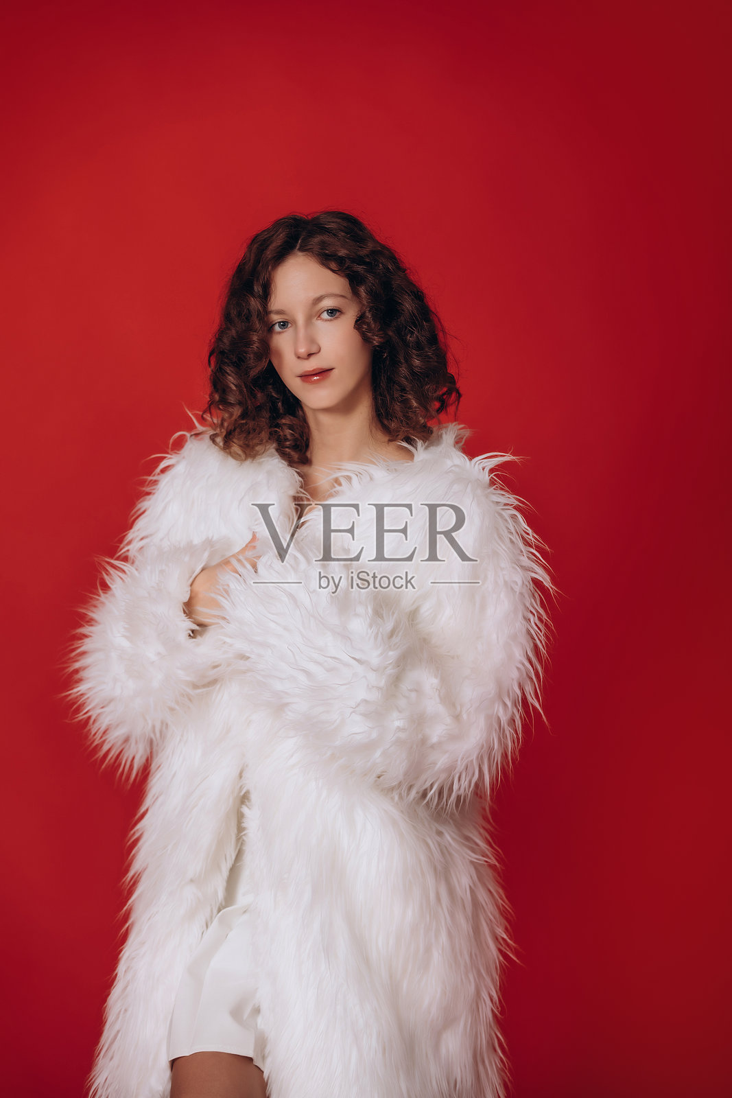 模特是一位年轻美丽的卷发棕发女孩，身穿白色蓬松皮草外套，背景为红色照片摄影图片