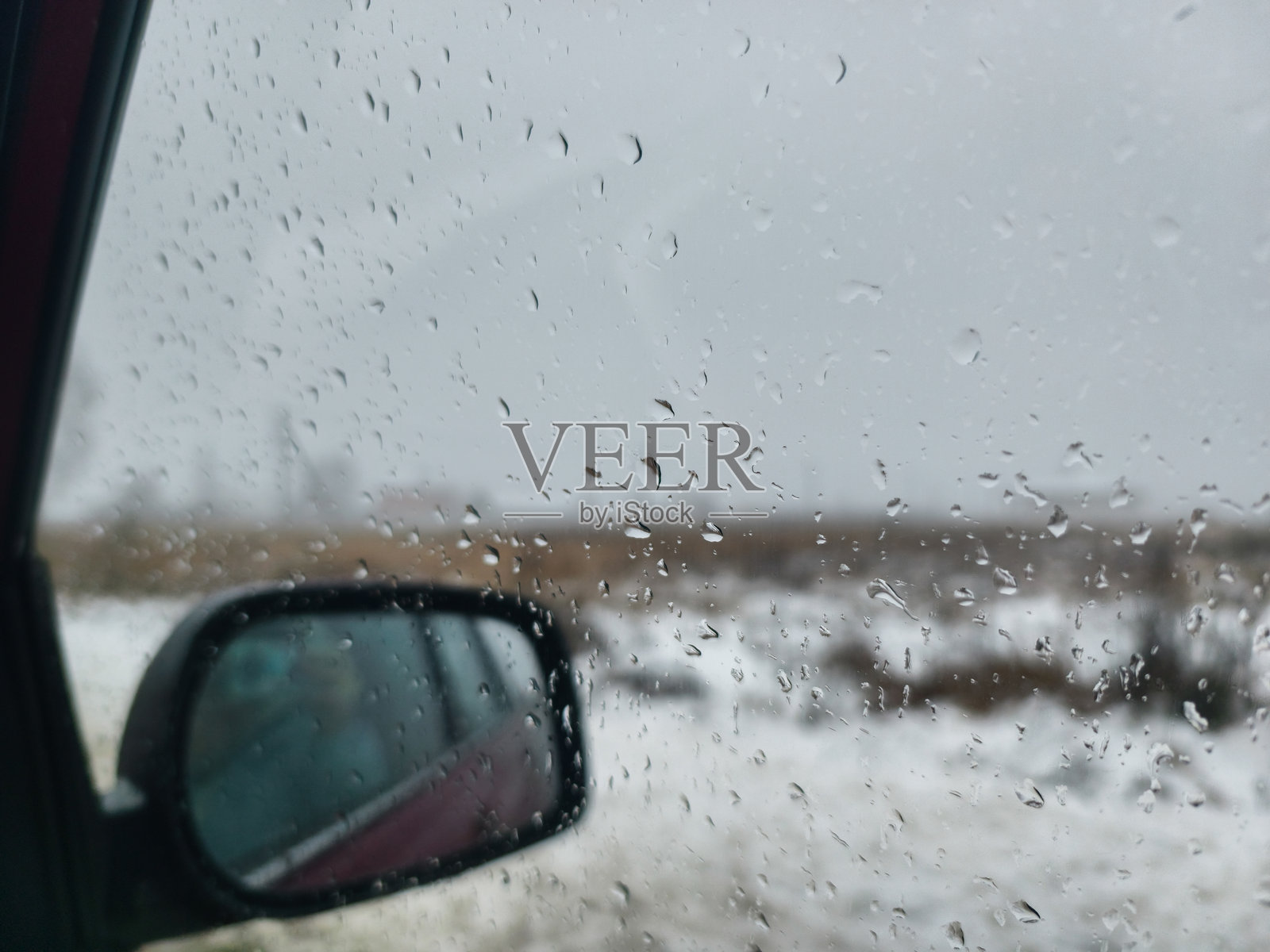 雨滴落在车窗上，模糊了窗外白雪皑皑的冬日景象。车子侧面的后视镜映照着灰色天空下的场景，营造出一种阴郁的氛围。照片摄影图片