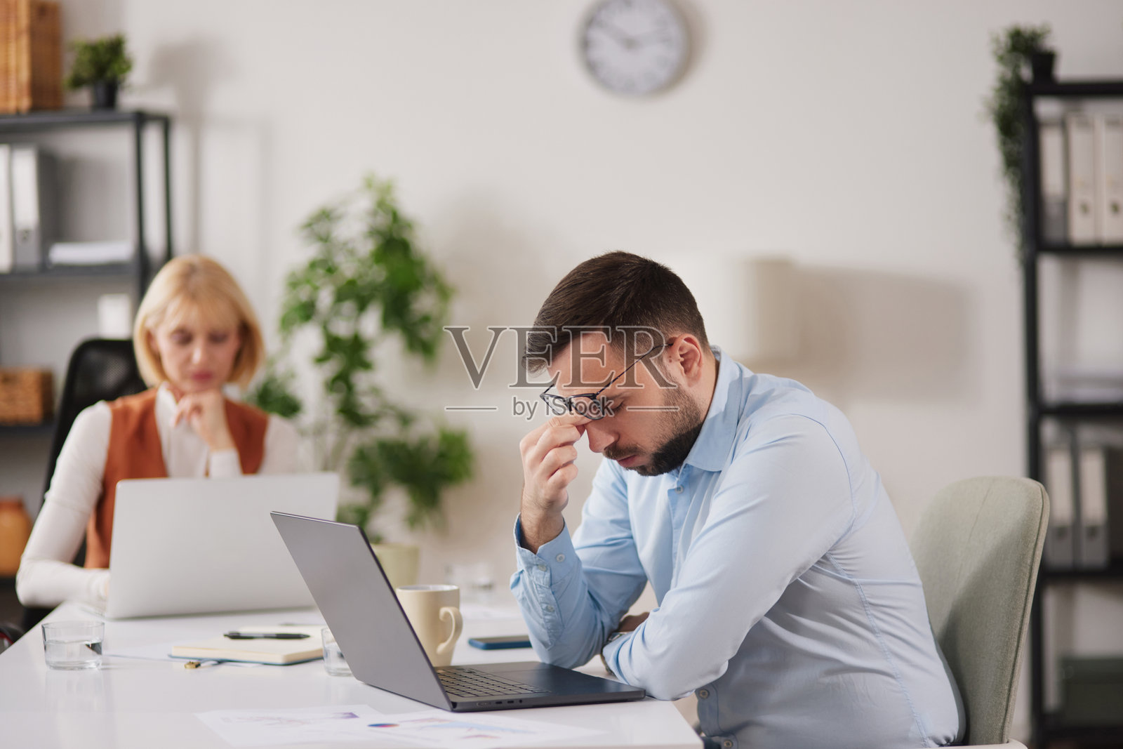 上班族在办公室用笔记本电脑工作时感到疲倦，背景有同事，是在繁忙的早晨工作场所。照片摄影图片