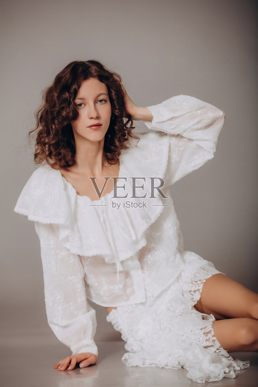 一位年轻美丽的卷发棕发女孩，身穿白色女衬衫和蕾丝裙，站在灰色背景前照片摄影图片