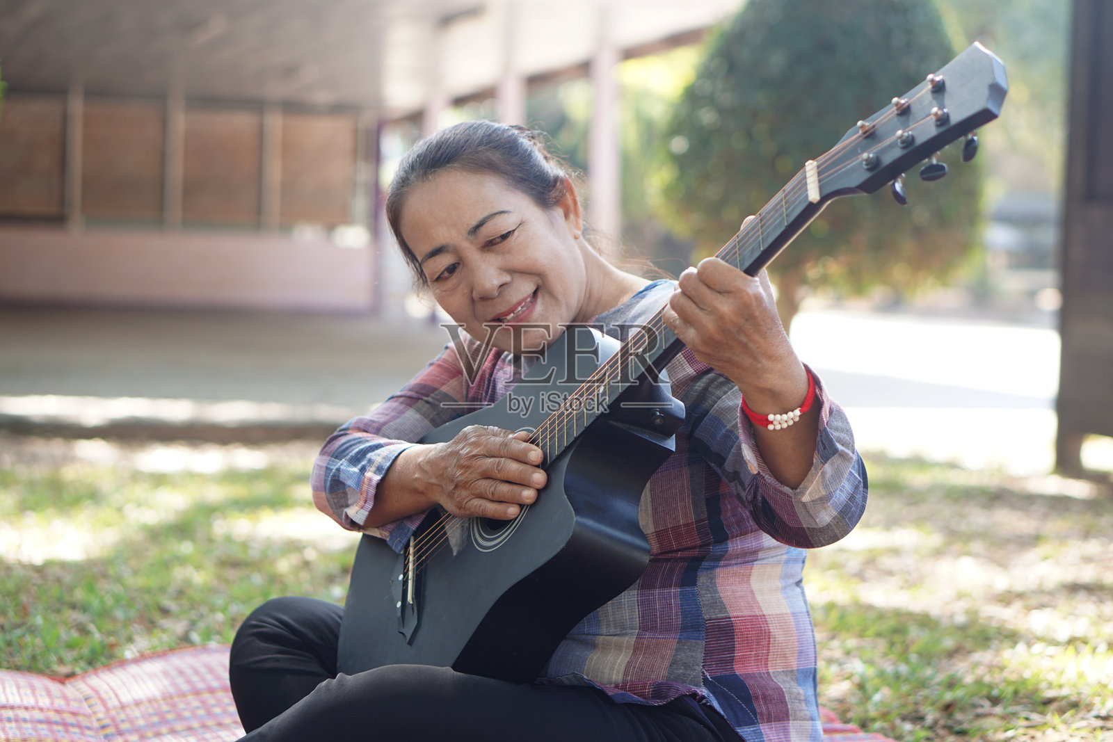 一位年长的亚洲女性在户外坐垫上弹奏木吉他。自信的女性享受音乐、创意爱好和健康老龄化生活方式，展现任何年龄的幸福和内在美。照片摄影图片