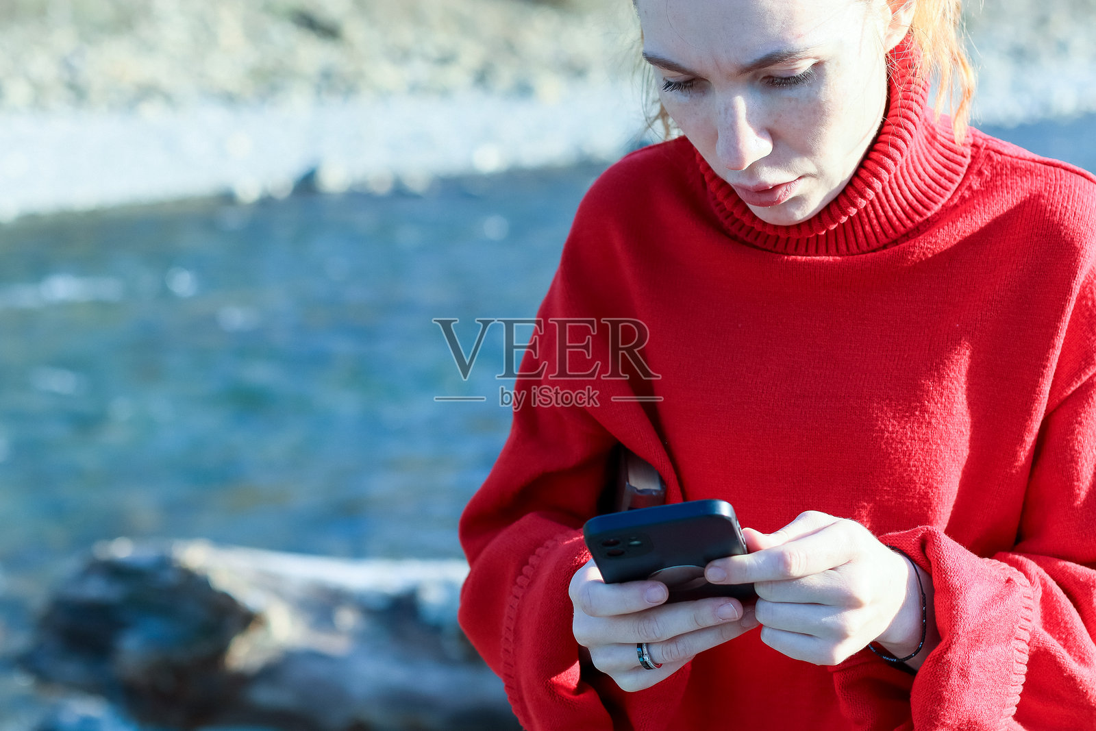 身穿红色高领毛衣的女子在河边使用智能手机，专注于屏幕，背景是自然风光。照片摄影图片