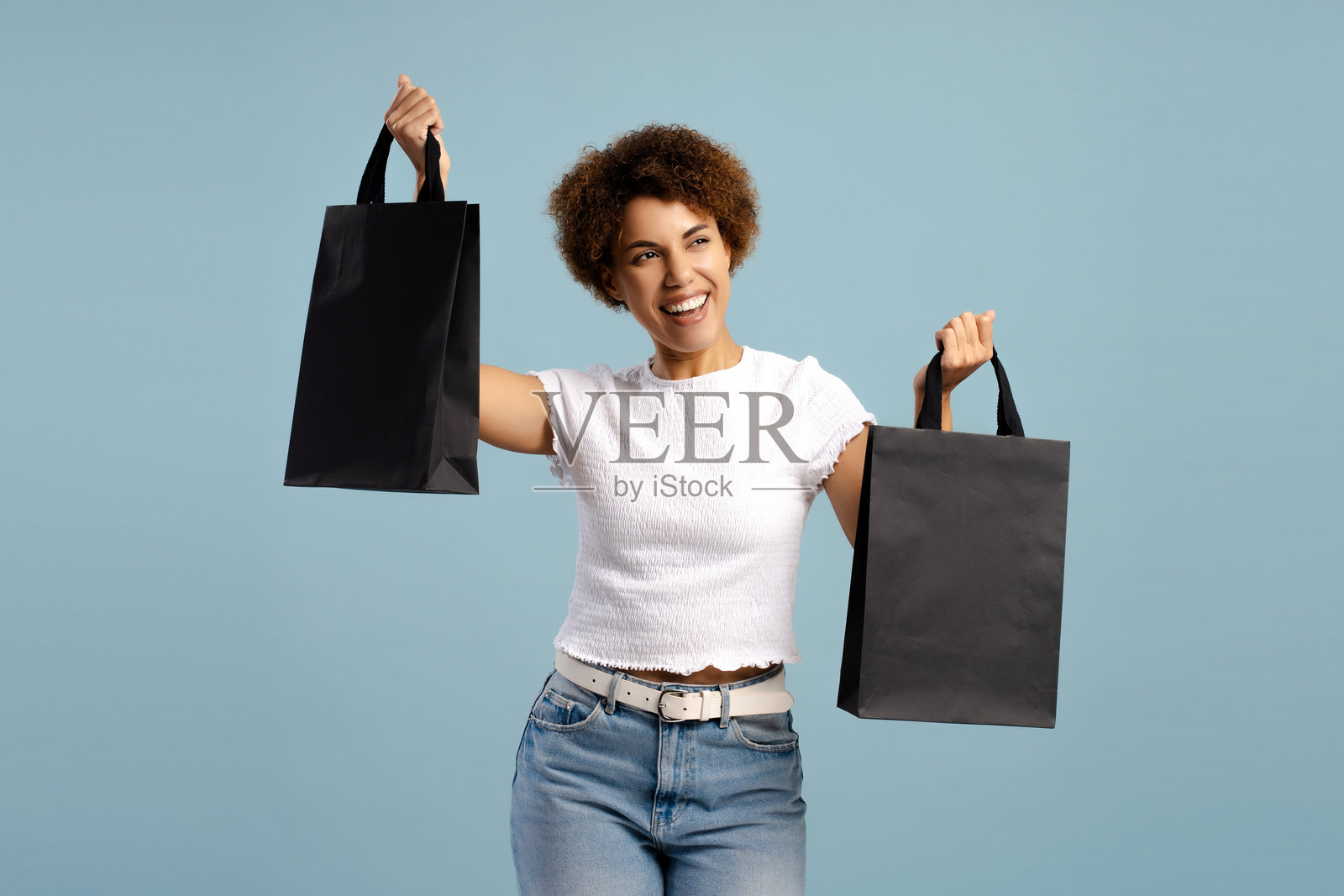 快乐的非裔美国女性，拎着黑色购物袋，享受购物的乐趣，在蓝色背景下被孤立。照片摄影图片