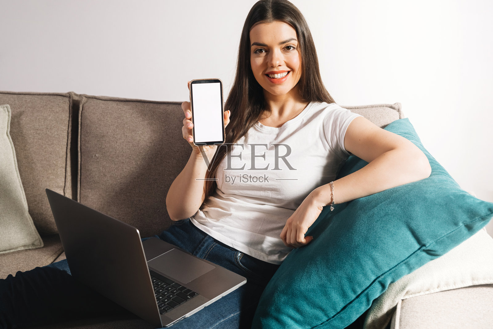 一位女士白天坐在沙发上，手持智能手机，旁边桌子上放着笔记本电脑，正在进行在线活动。照片摄影图片