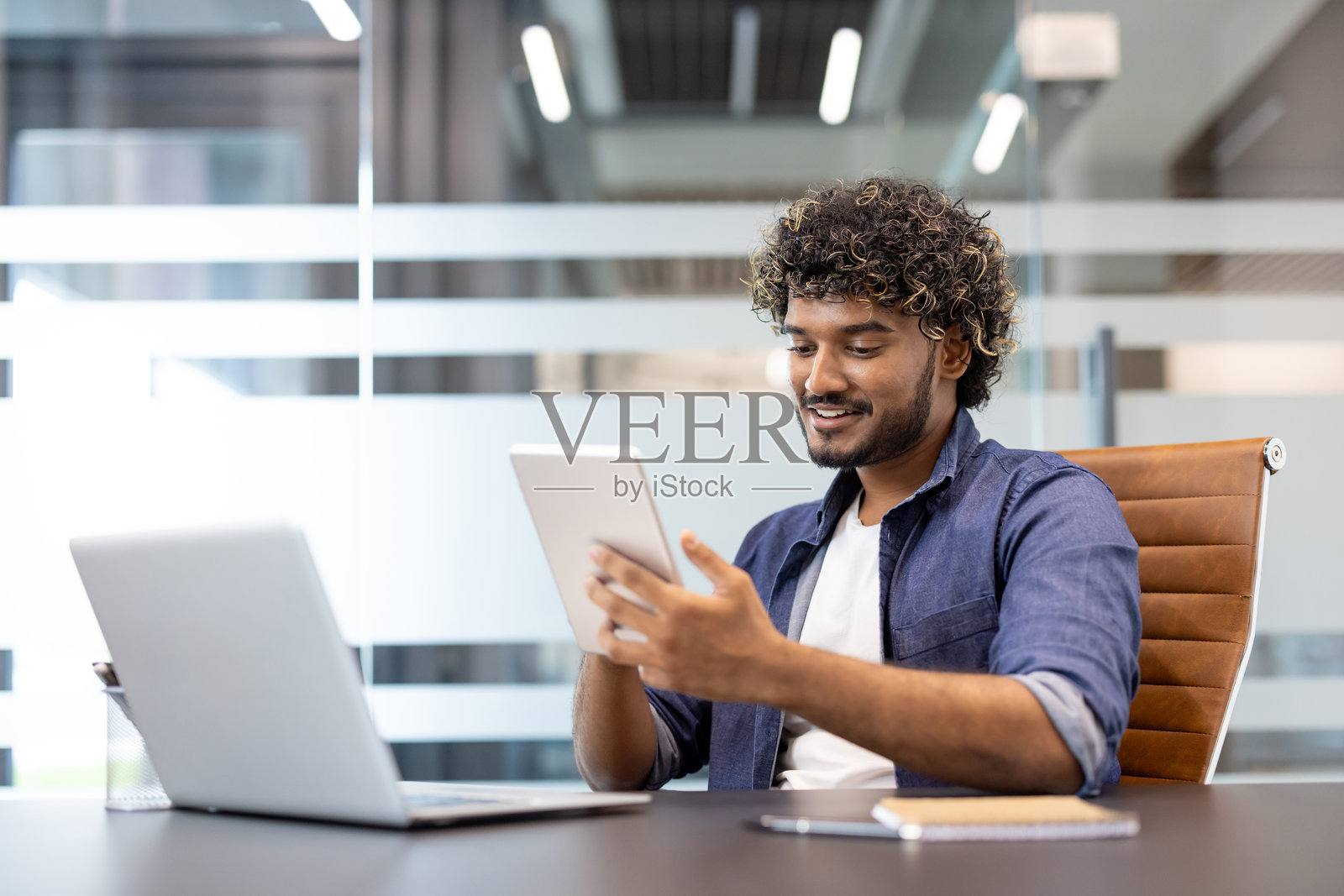 一位年轻的印度男子坐在办公室的桌子前，使用平板电脑工作照片摄影图片