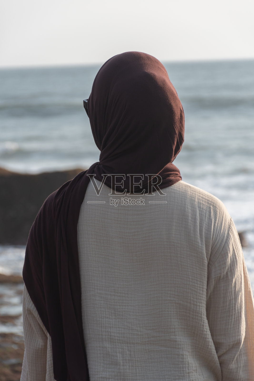 一位戴着头巾的女性站在海边，若有所思地凝视着地平线。照片摄影图片