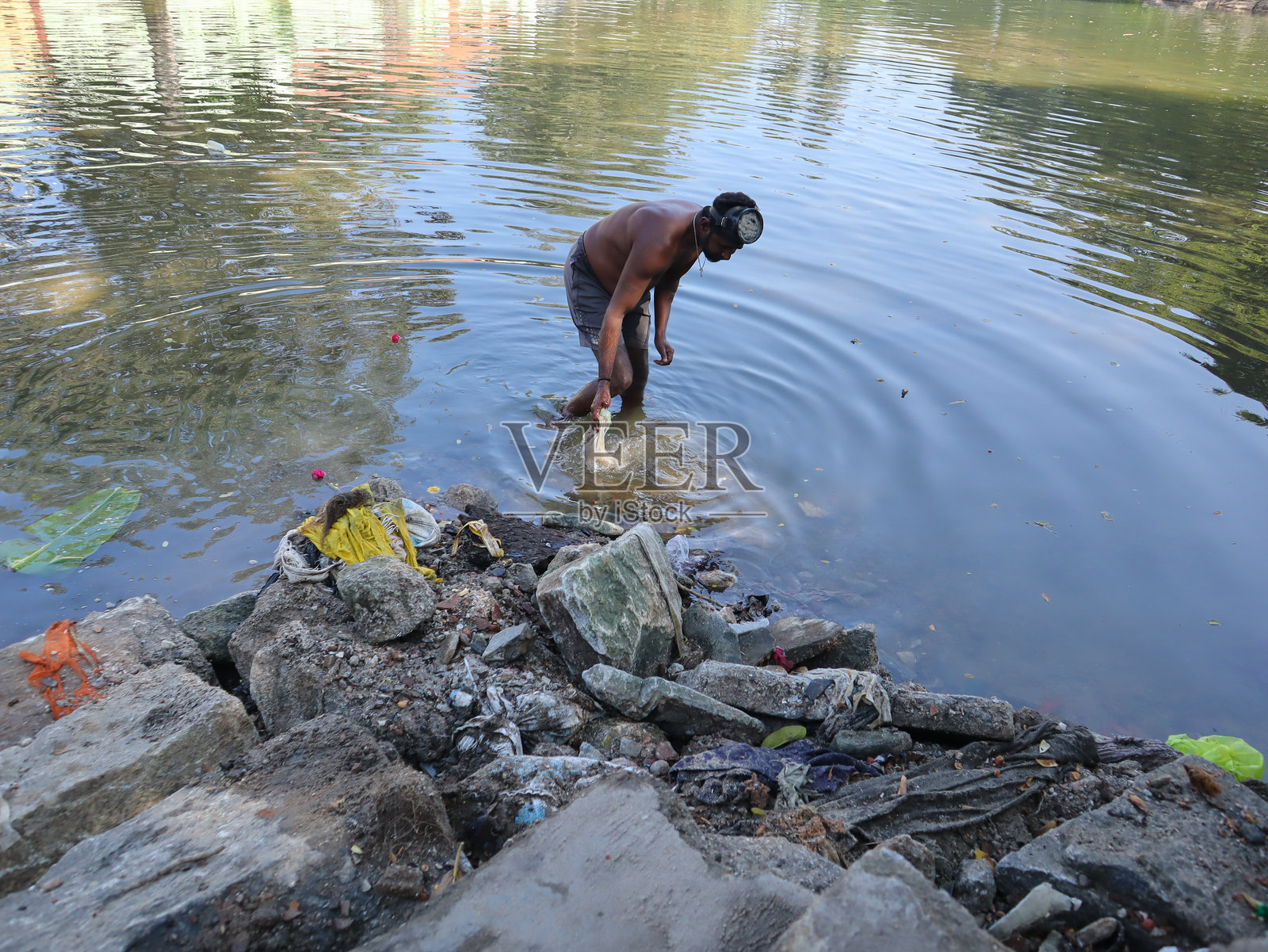 印度卡纳塔克邦，一名市政工人正在打捞被污染的河水中的垃圾、塑料、衣物和淤泥，以进行废物管理和环境保护。照片摄影图片