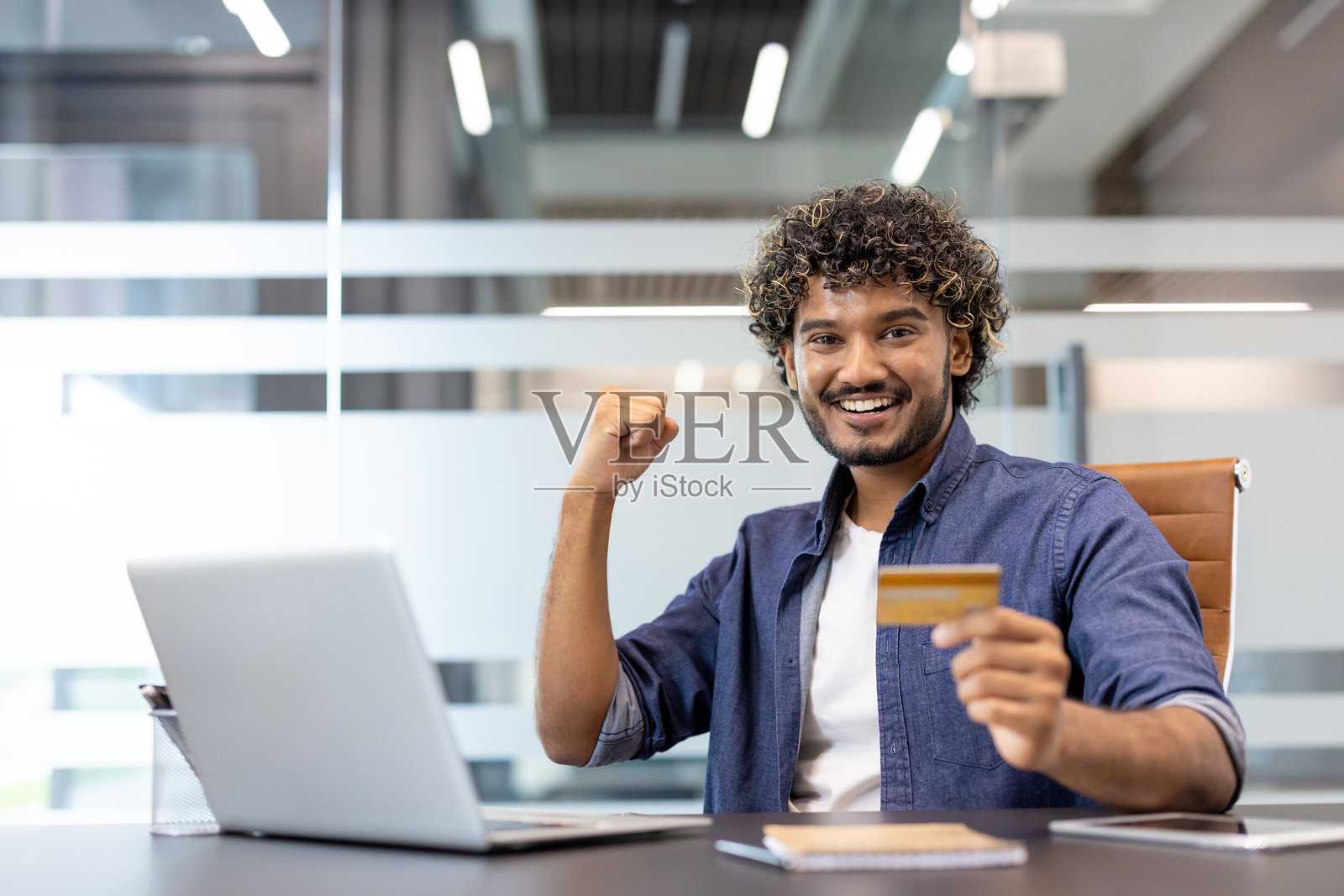 一位年轻快乐的印度男子在办公室的桌子前，坐在笔记本电脑前，手中拿着信用卡，为成功而欢欣鼓舞照片摄影图片