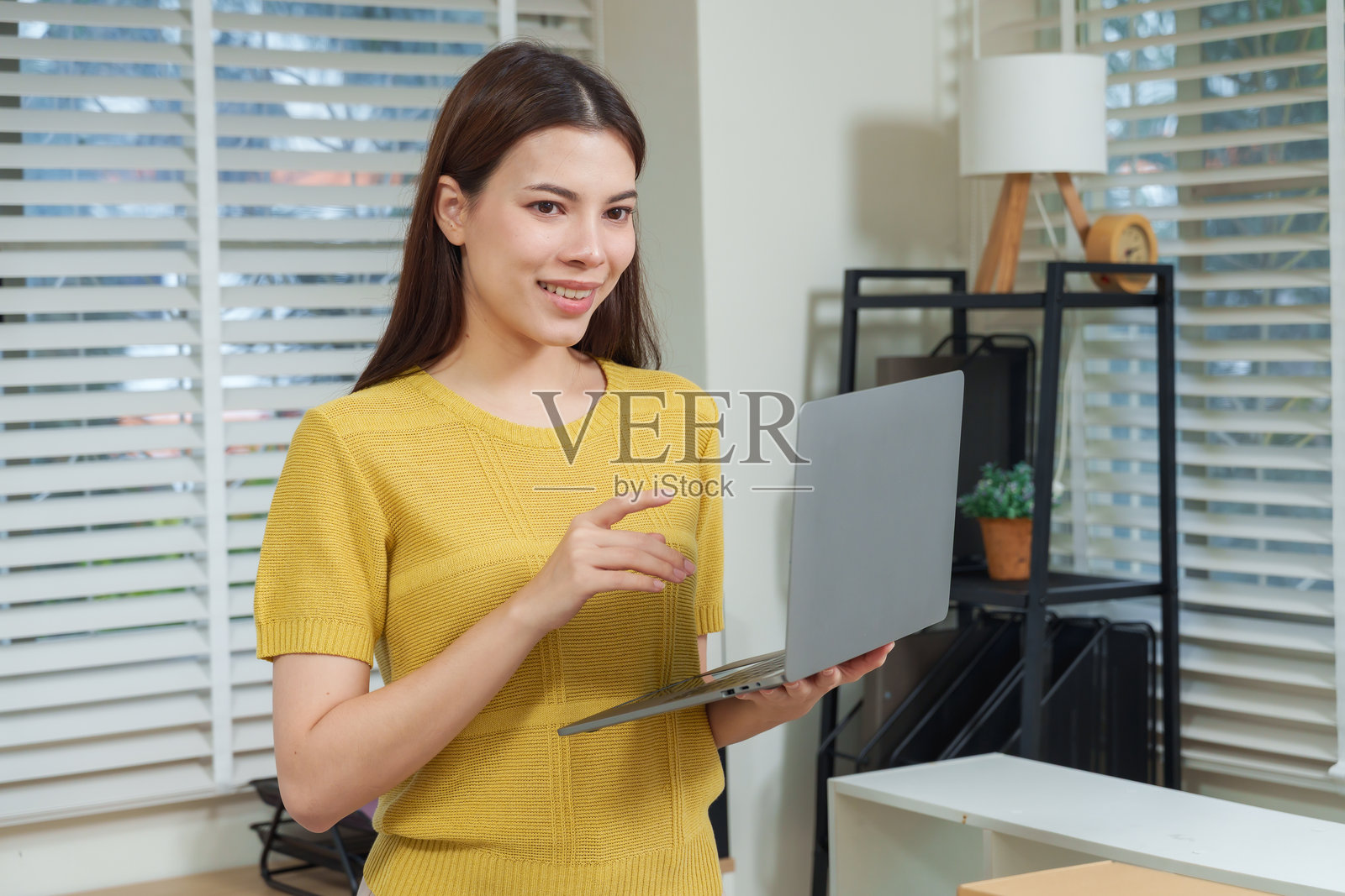 亚洲女商人手持笔记本电脑，面带微笑，与客户在线交流，体现了明亮现代办公室中的职业成功、数字沟通和创业精神的概念。照片摄影图片