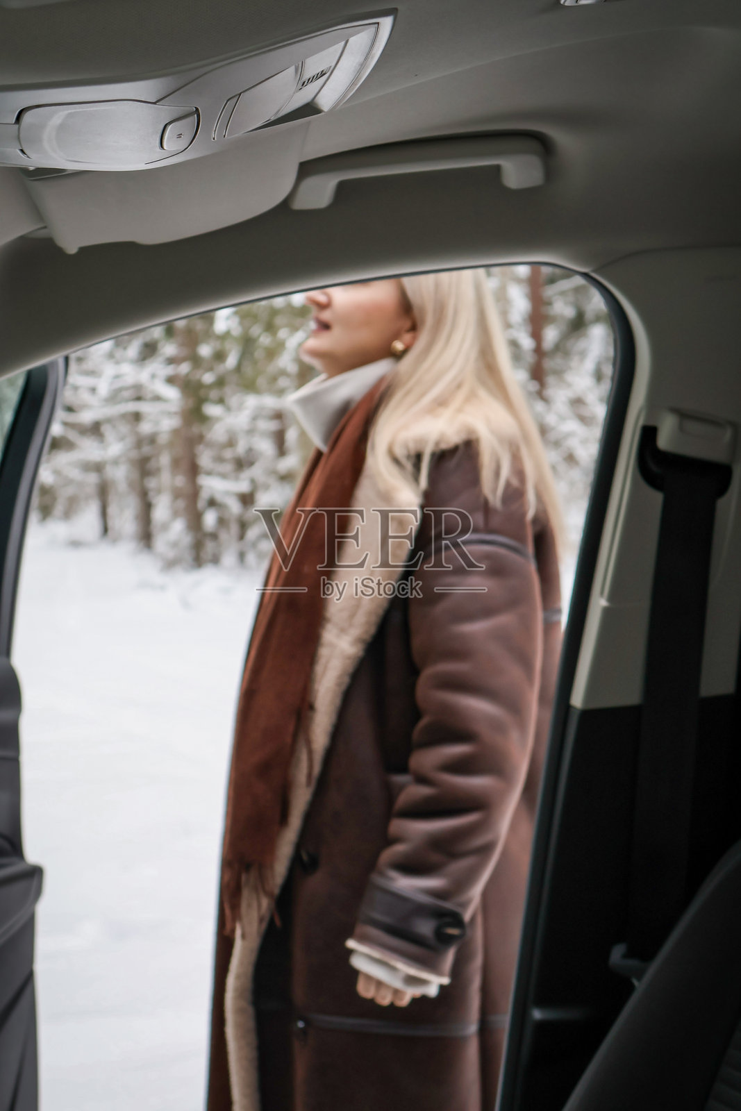 从车内望去，一位中年女性穿着暖和的外套，站在雪林旁的汽车旁。照片摄影图片