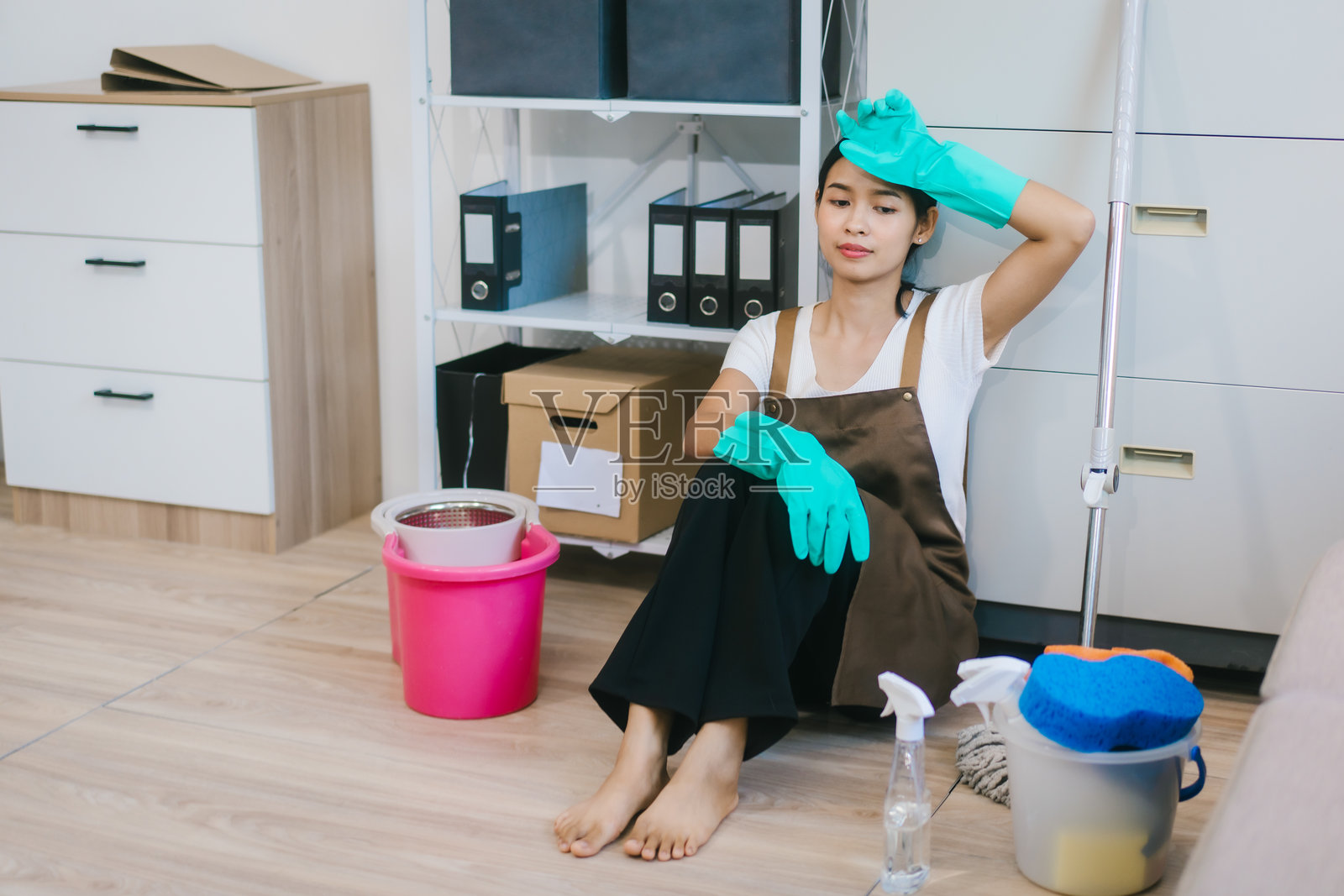 一位穿着棕色围裙的亚洲女性清洁工坐在木地板上，在短暂休息时微笑着接听电话，周围是整洁工作空间里的清洁用品。照片摄影图片