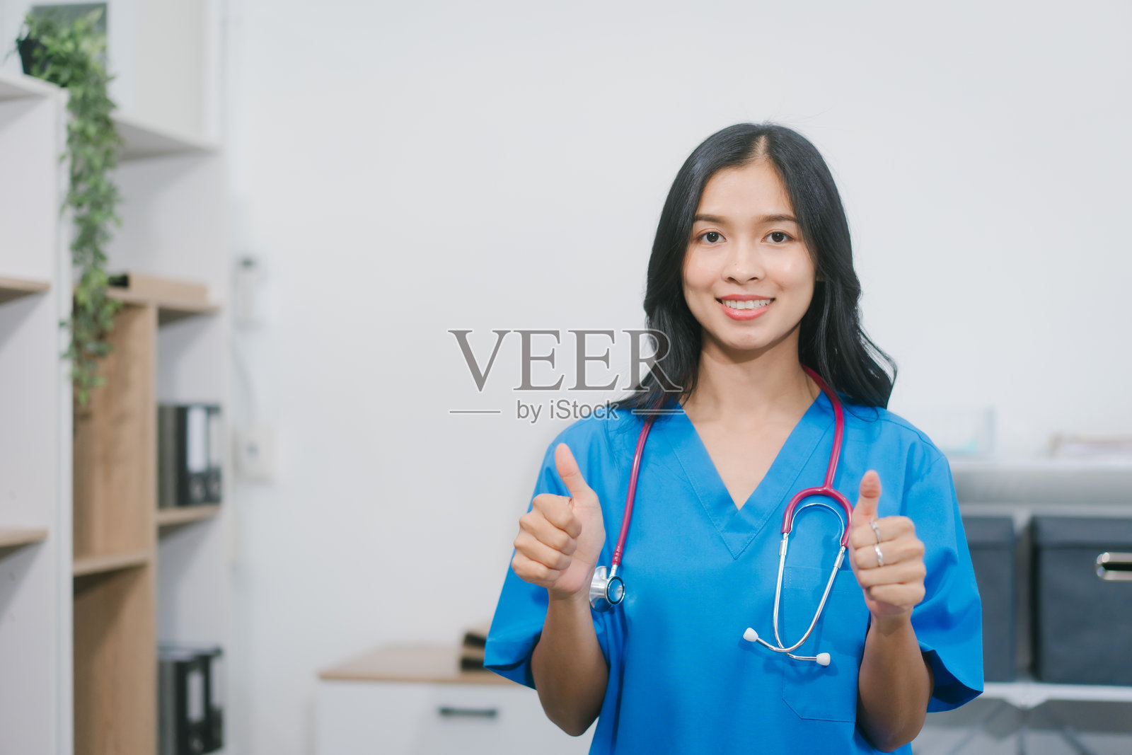 一位身穿蓝色工作服的亚洲女护士自信地站在明亮的诊室里，手持病历表和听诊器，笑容温暖，代表着医疗的专业与信赖。照片摄影图片