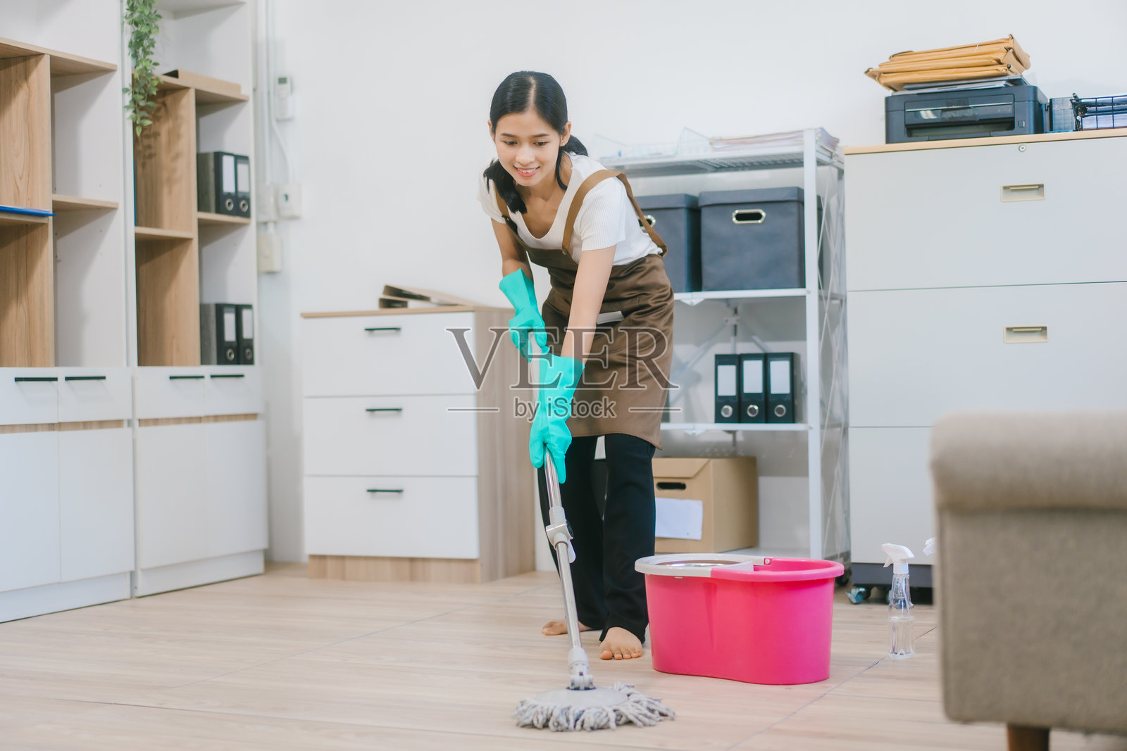 一位戴着手套和围裙的亚洲女性家政服务人员正在清洁现代办公室或家居。她勤奋工作，维护卫生和秩序，体现了专业的清洁服务。照片摄影图片