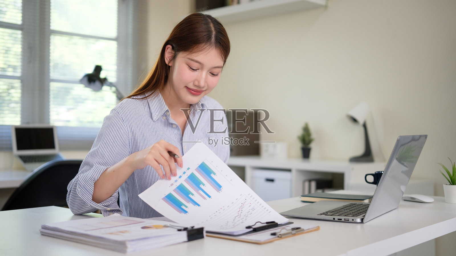 年轻的职业女性在办公室的办公桌上用笔记本电脑工作并审阅文件。照片摄影图片