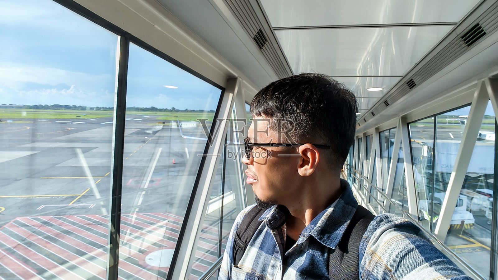 一个穿着休闲衬衫、背着背包的男人，在一个阳光明媚的下午，走在机场的廊桥上，望着窗外。照片摄影图片