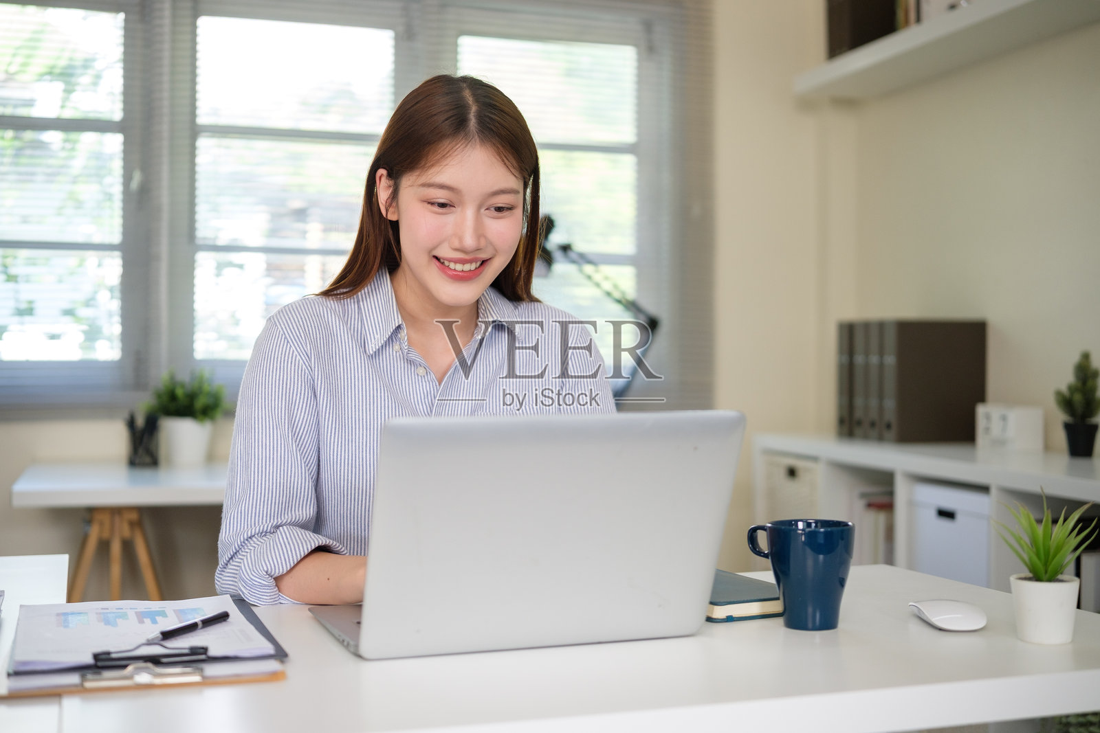 年轻的商界女性在现代办公室的办公桌前，微笑着使用笔记本电脑处理财务文件。照片摄影图片