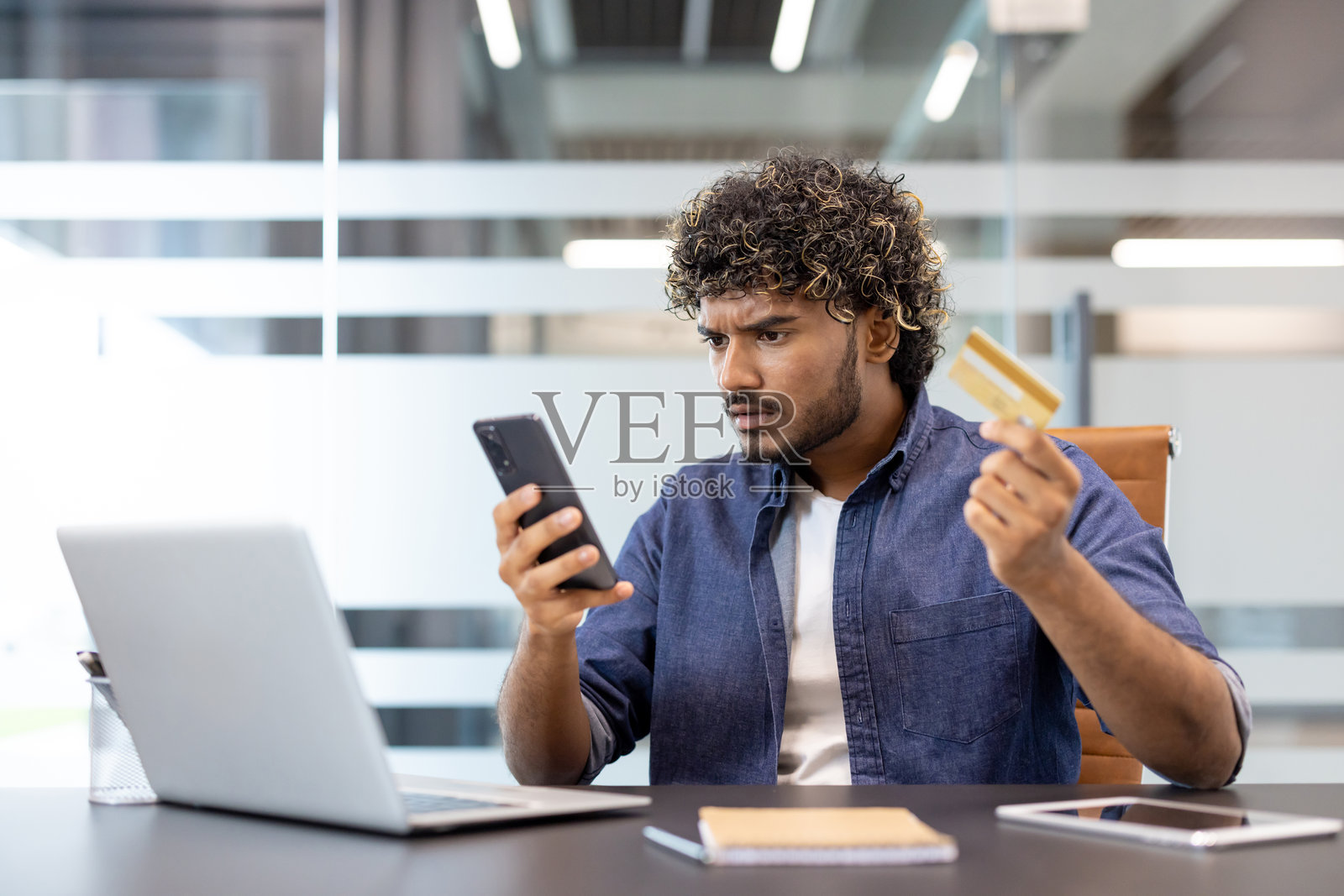 一位年轻的印度男子，在办公室里坐在桌前，手里拿着信用卡，沮丧地看着手机。照片摄影图片