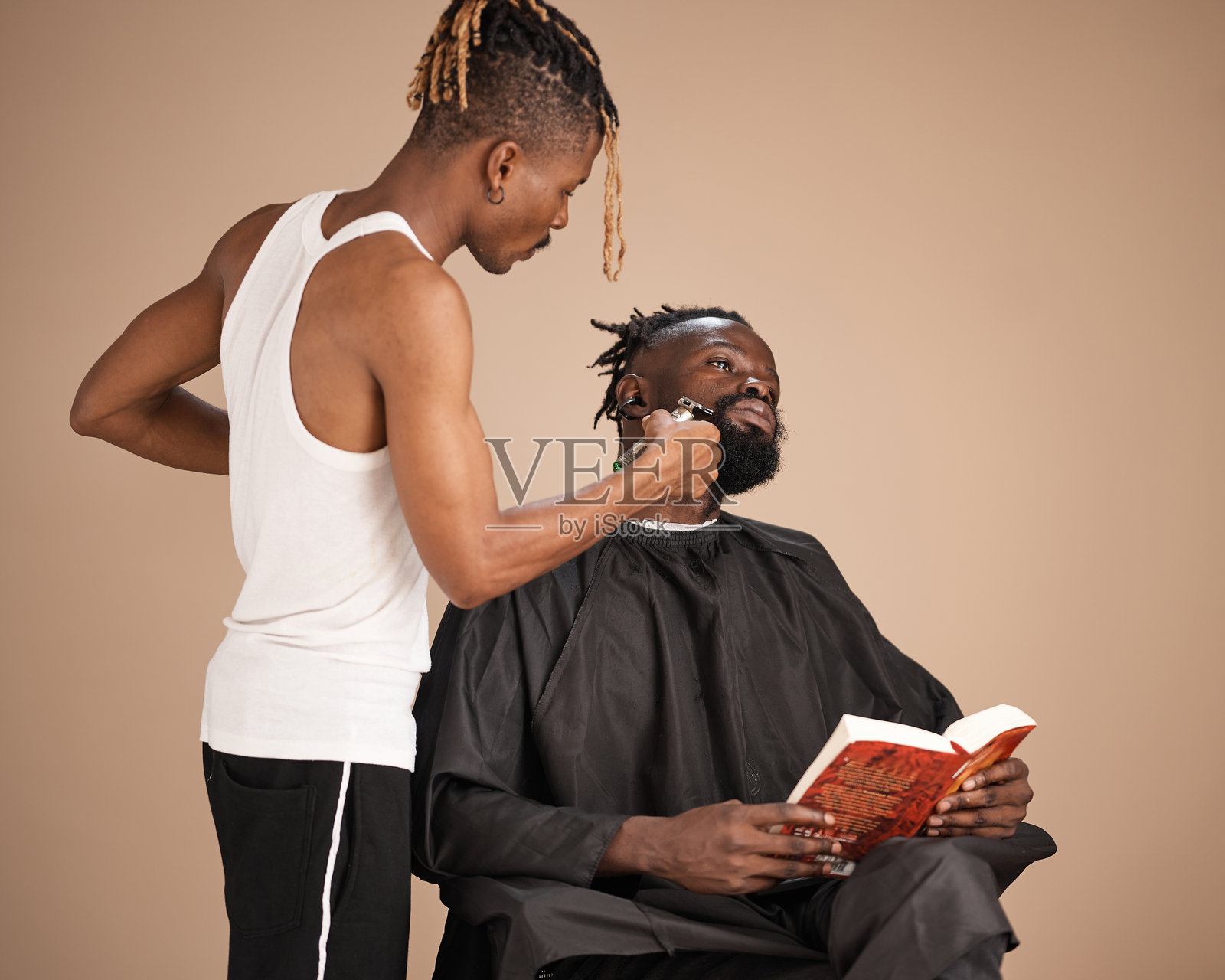 年轻的非洲理发师在工作室里为顾客修剪胡须，顾客在一旁看书照片摄影图片