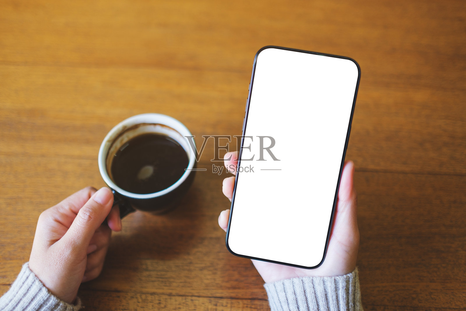 俯视角度的样机图片，展示了一位女士一手拿着手机，另一手端着咖啡，手机屏幕为空白桌面。照片摄影图片