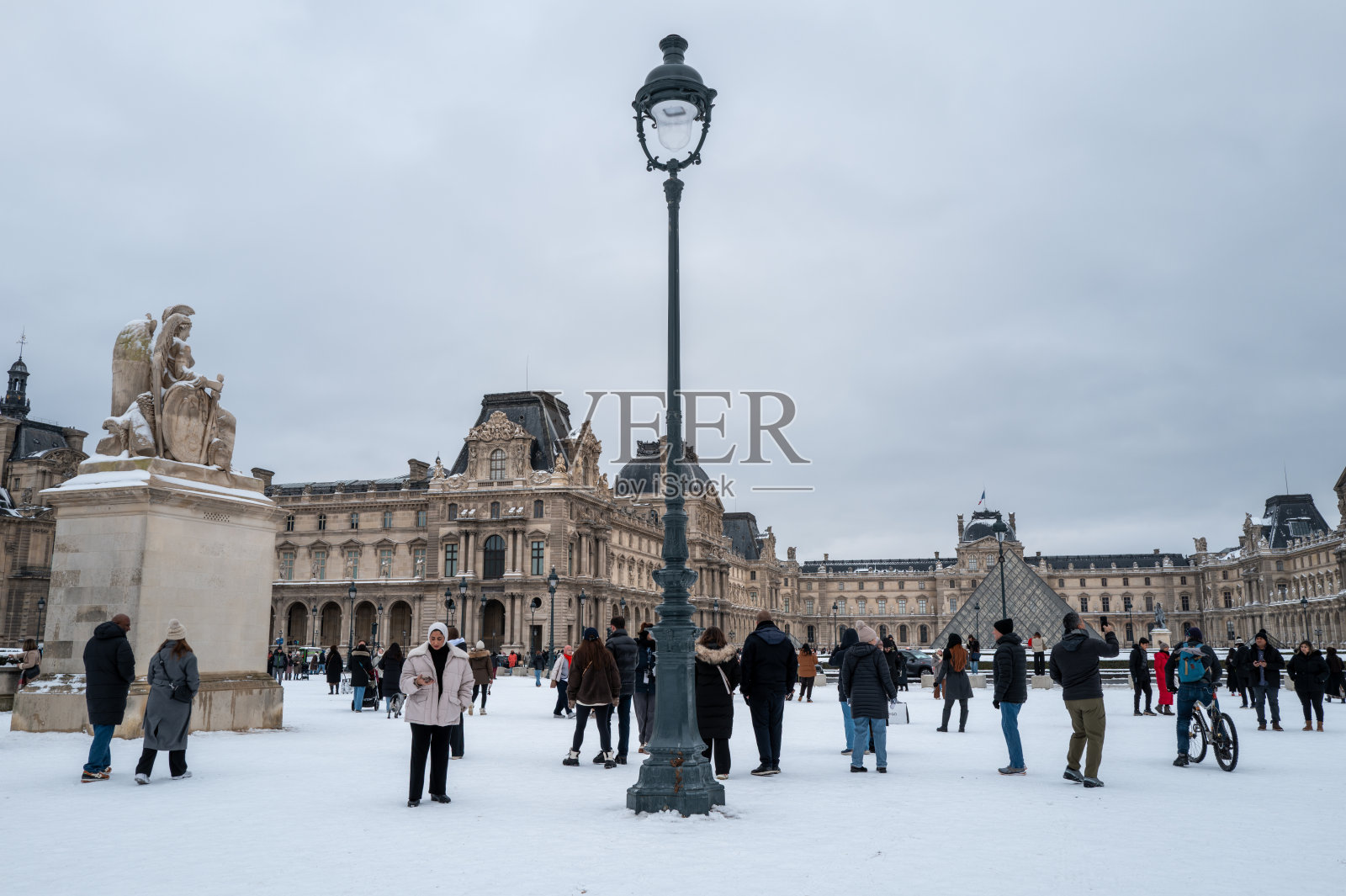 法国巴黎卢浮宫博物馆内，人们在被白雪覆盖的庭院中，背景是玻璃金字塔和雕像。照片摄影图片