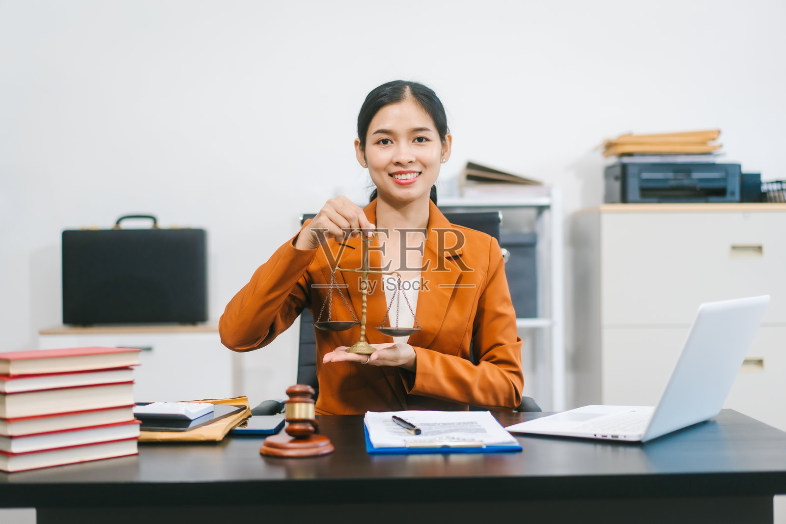 一位年轻的亚洲女性身着正式西装外套，在现代办公室自信地使用笔记本电脑，周围散落着法律文件和专业工具，展现出专业、专注和精致的企业形象。照片摄影图片