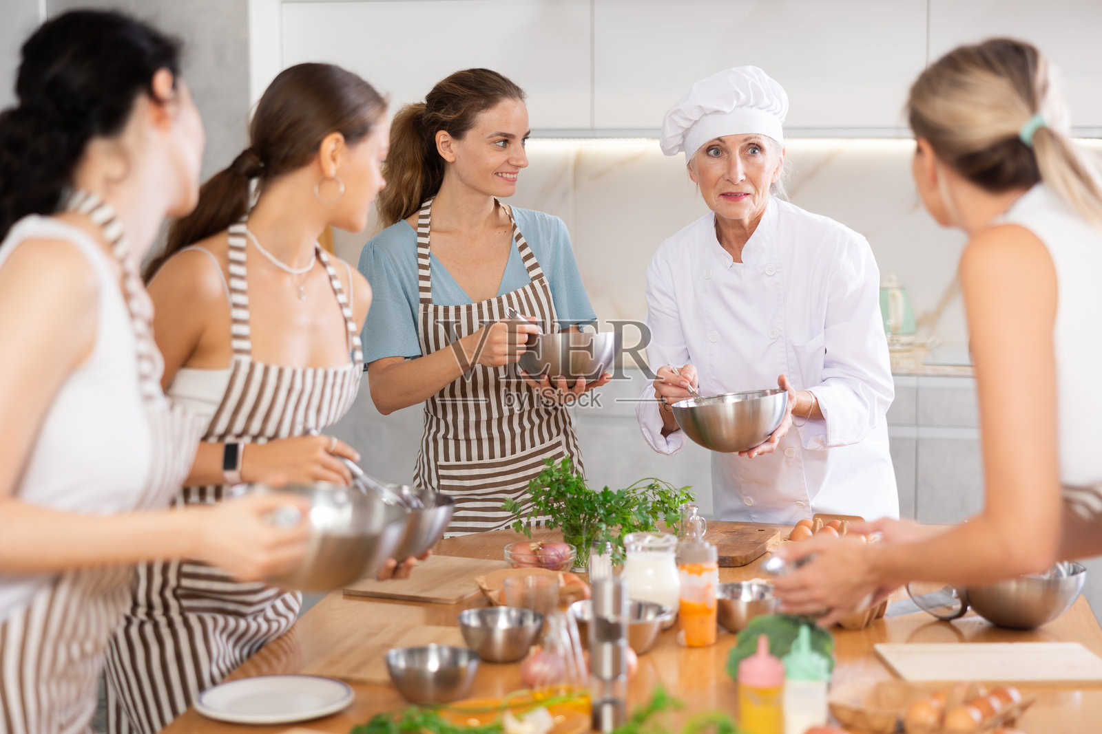 一位年长的女性厨师，手捧着碗，在烹饪课程上向女性们讲述美食传统。照片摄影图片