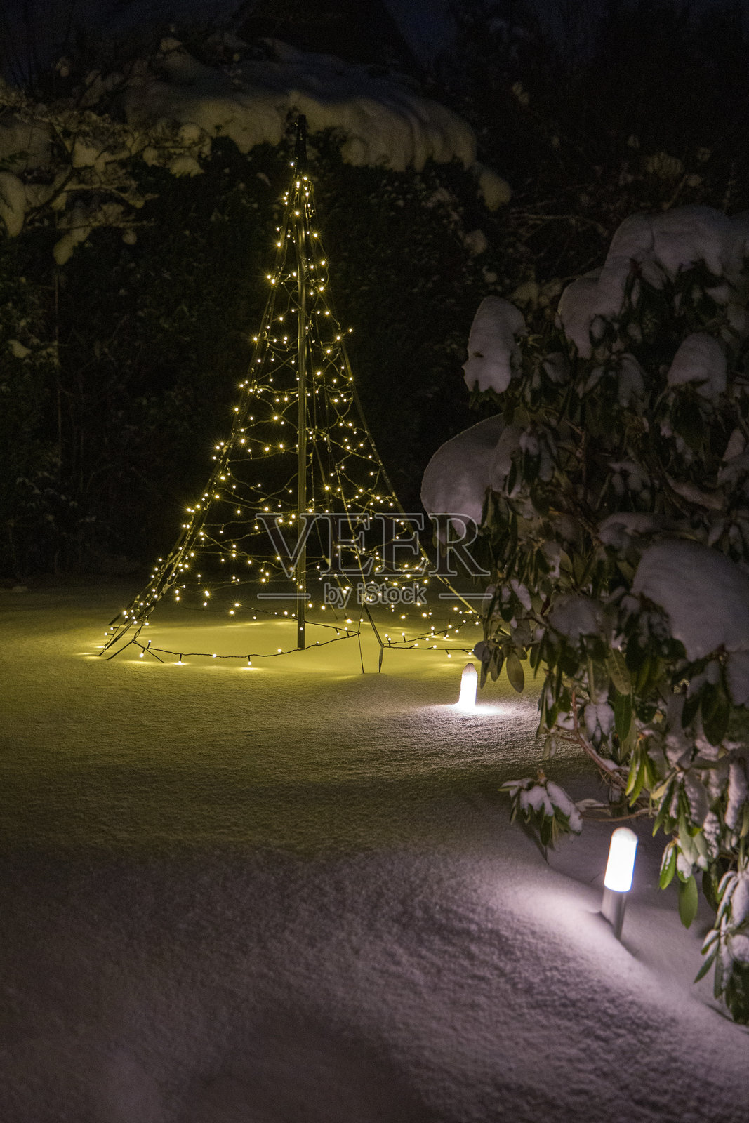 夜晚，雪覆盖的花园里，一棵由灯链和花园灯组成的发光圣诞树照片摄影图片