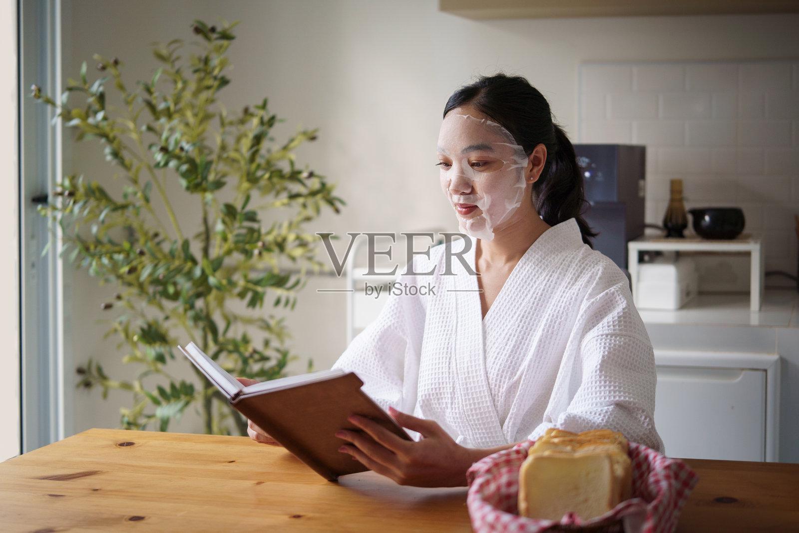 一位女性在舒适的早晨例行项目中，戴着面膜在厨房的餐桌旁读书。照片摄影图片