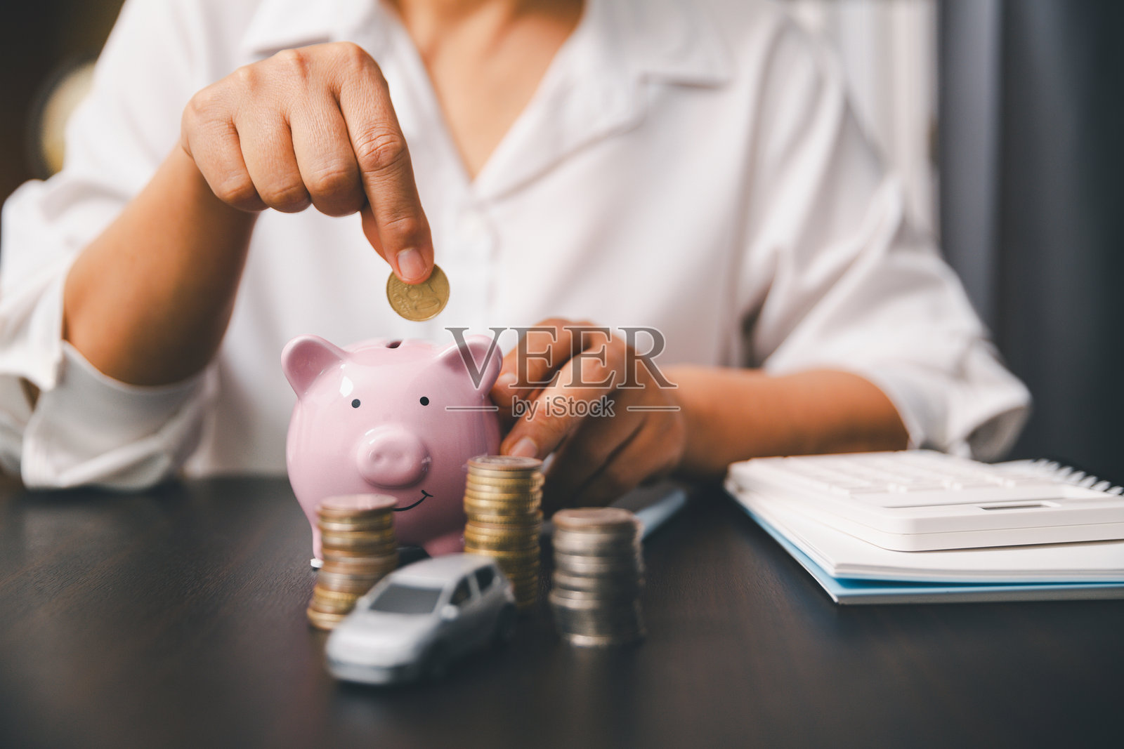 手将硬币投入粉色储蓄罐，旁边桌子上放着钱币堆、玩具车和计算器，展示了汽车储蓄、首付、汽车贷款预算、保险费用和财务规划。照片摄影图片