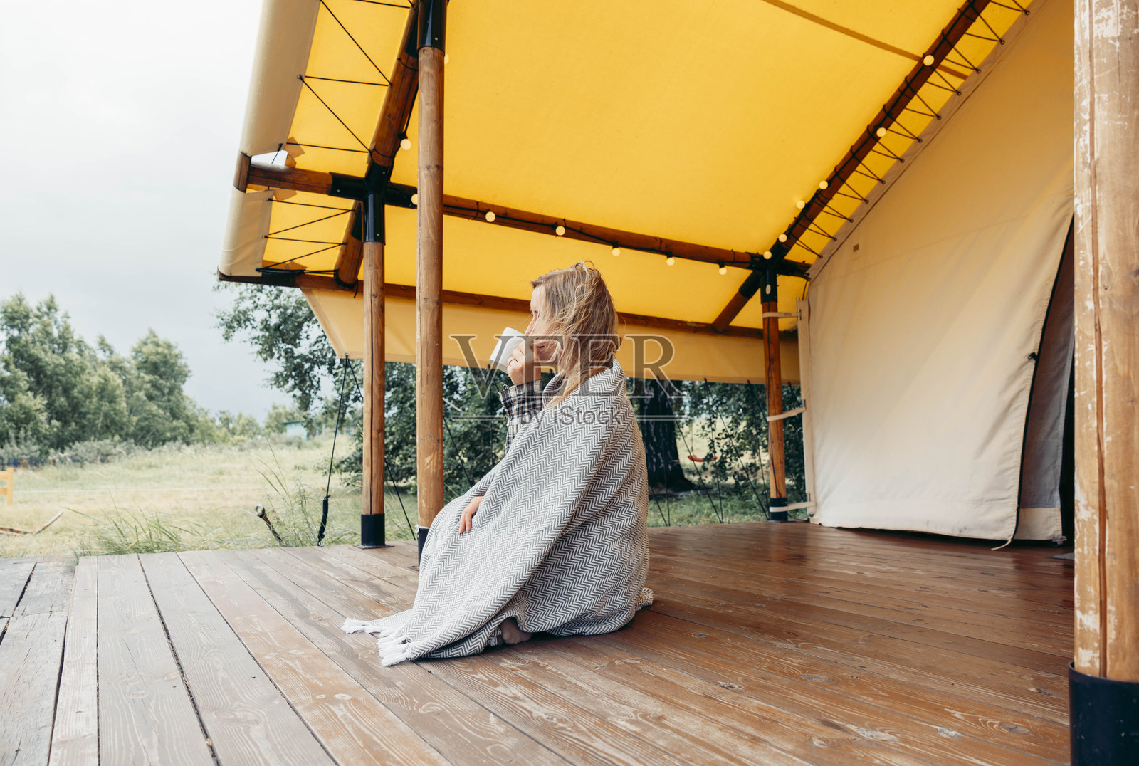 一位女子裹着毯子，坐在露营帐篷的木质露台上，手持白色马克杯，在宁静的乡村早晨凝视远方，享受露营和旅行的生活方式照片摄影图片