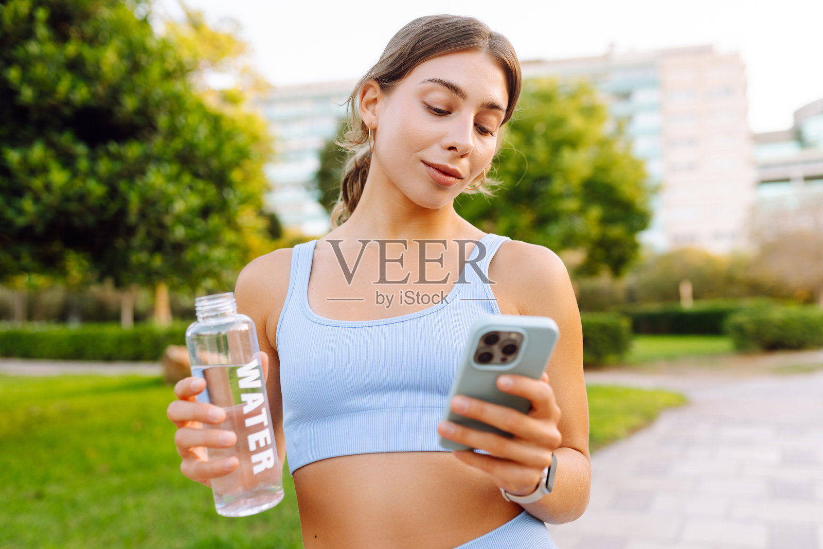 身穿运动服的快乐女子手持手机和一瓶水，站在阳光明媚的公园里。照片摄影图片