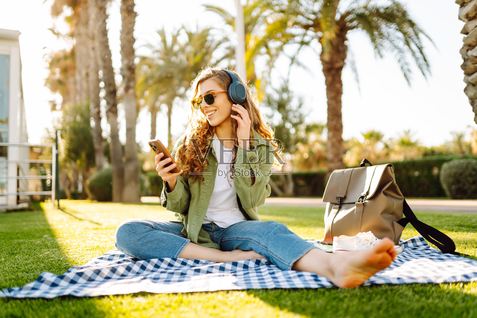 年轻女子戴着无线耳机，在阳光明媚的公园里，周围环绕着棕榈树，她正看着手机。照片摄影图片