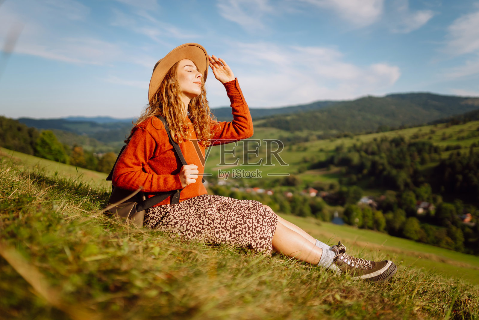 戴着帽子、背着背包的年轻女子在绿色的山坡上享受阳光明媚的一天。照片摄影图片