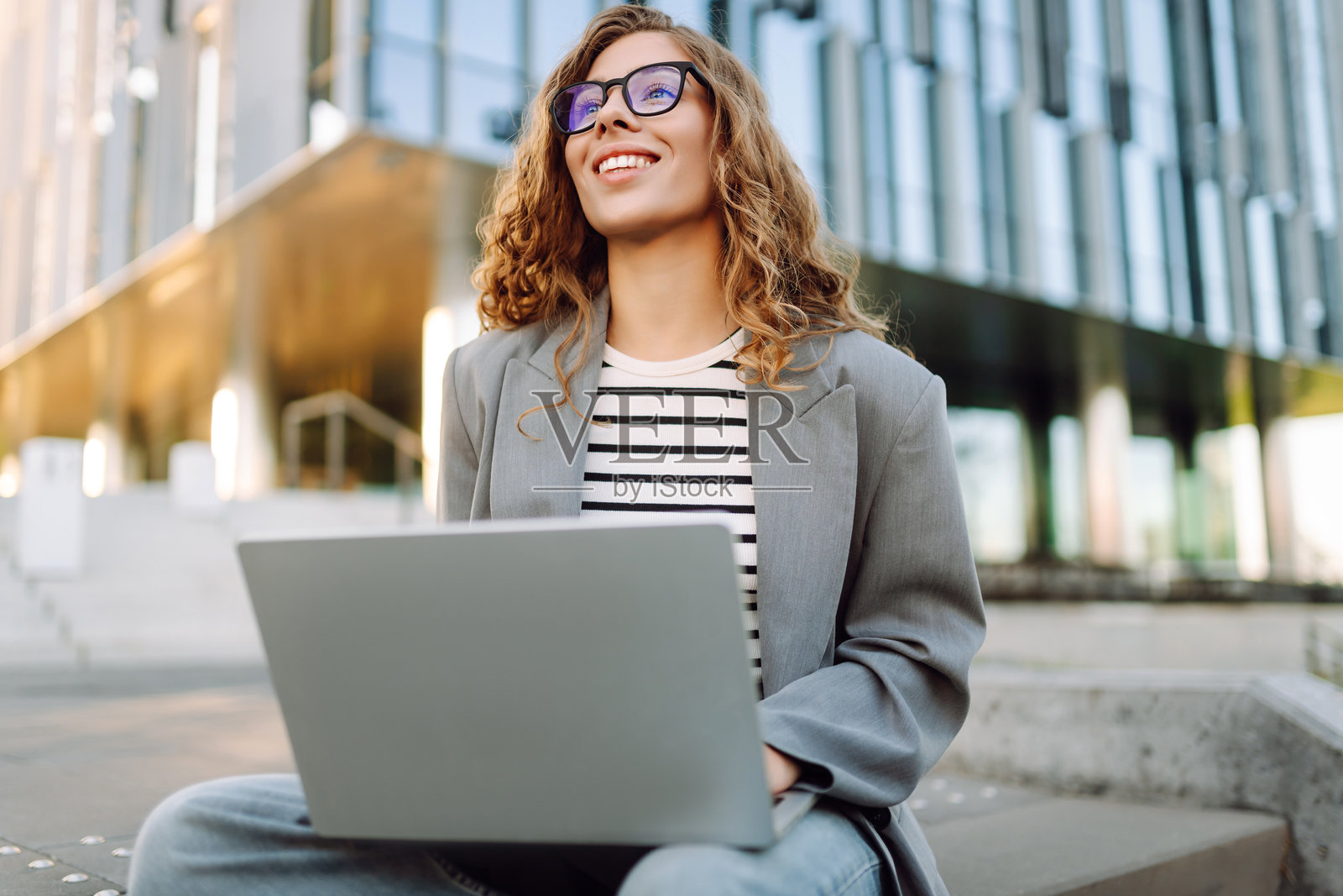 一位时尚女子坐在台阶上，背景为现代建筑，她正使用笔记本电脑工作。照片摄影图片