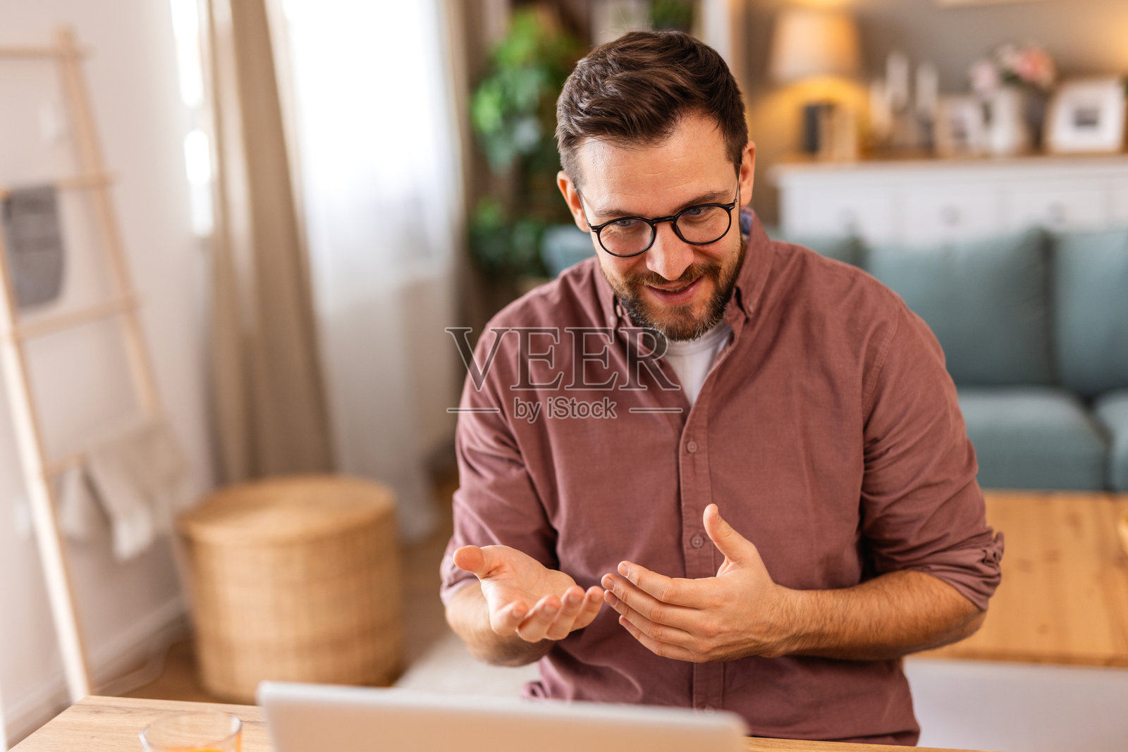 戴眼镜的微笑男士在家中办公室的笔记本电脑上进行视频通话时比划着，一位专业的男性自由职业者在客厅进行在线会议或远程工作网络研讨会时解释项目。照片摄影图片