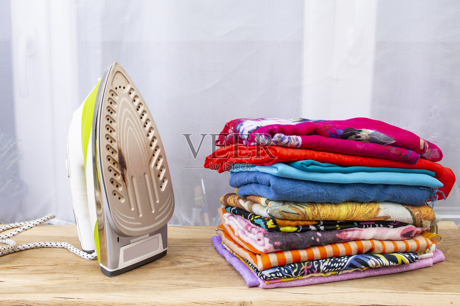 木桌上，带有烧焦痕迹的熨斗旁边堆放着一叠色彩鲜艳的折叠衣物或围巾照片摄影图片