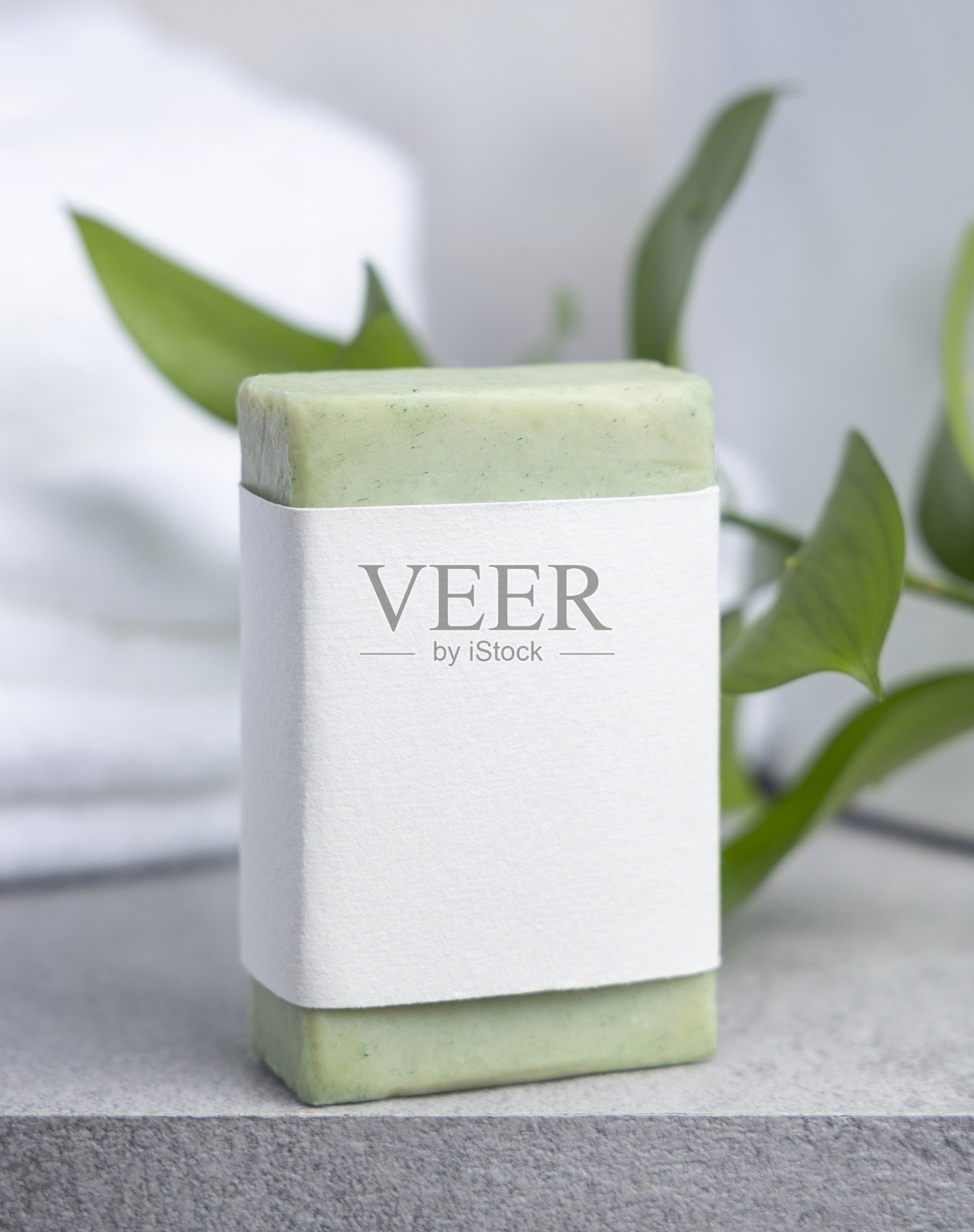 带有空白标签的肥皂块，背景为绿叶和白色浴巾，置于灰色石头上，样机照片摄影图片